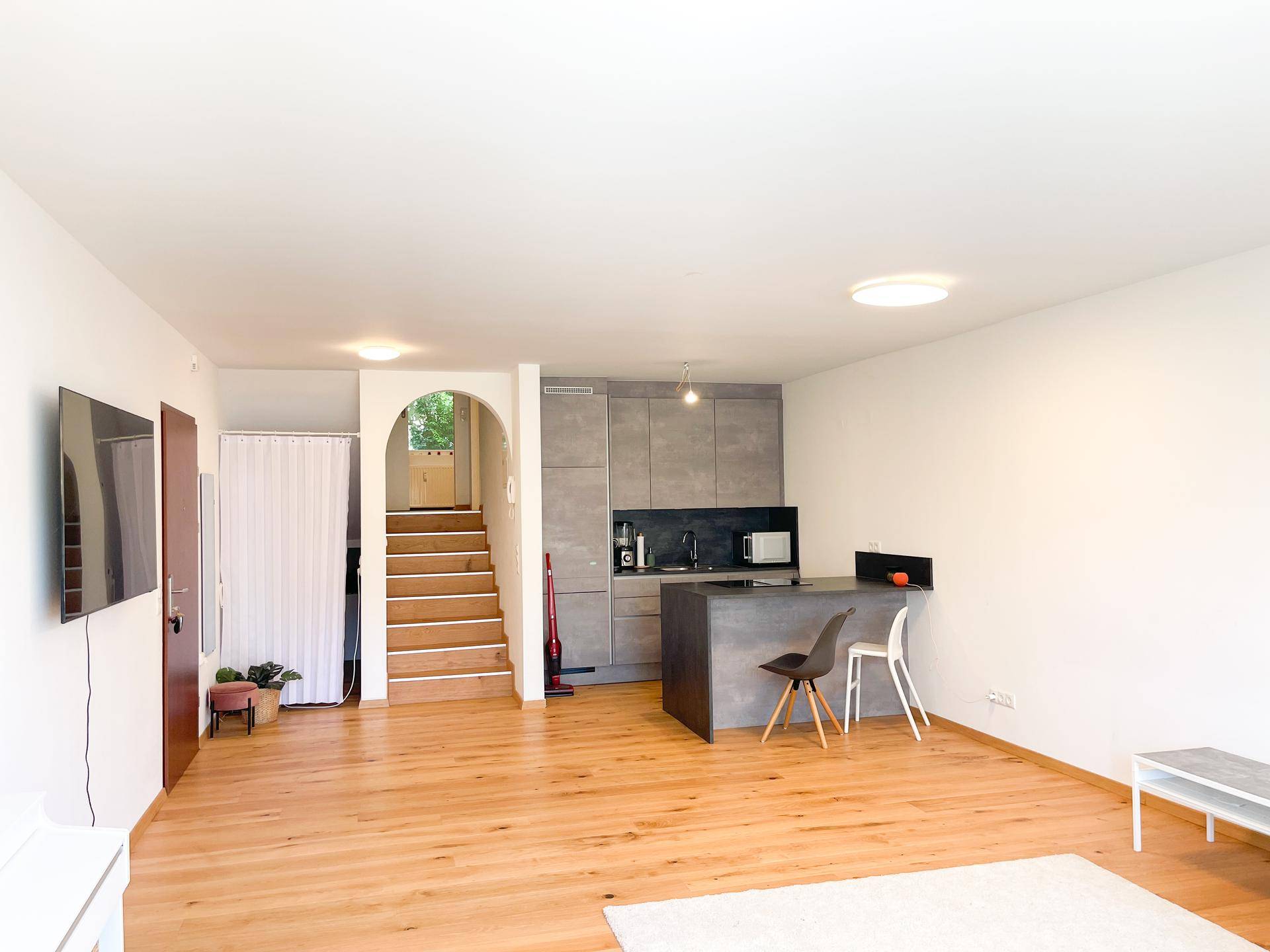 Offener Wohnbereich mit integrierter Küche, Holzboden und Zugang zum oberen Stockwerk über eine Treppe.