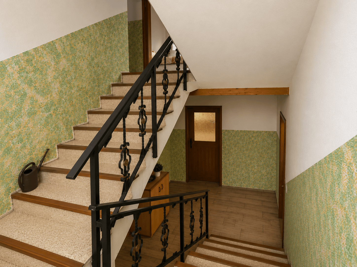 Treppenhaus mit gemusterter Tapete, Holzgeländer und verschiedenen Bodenbelägen.