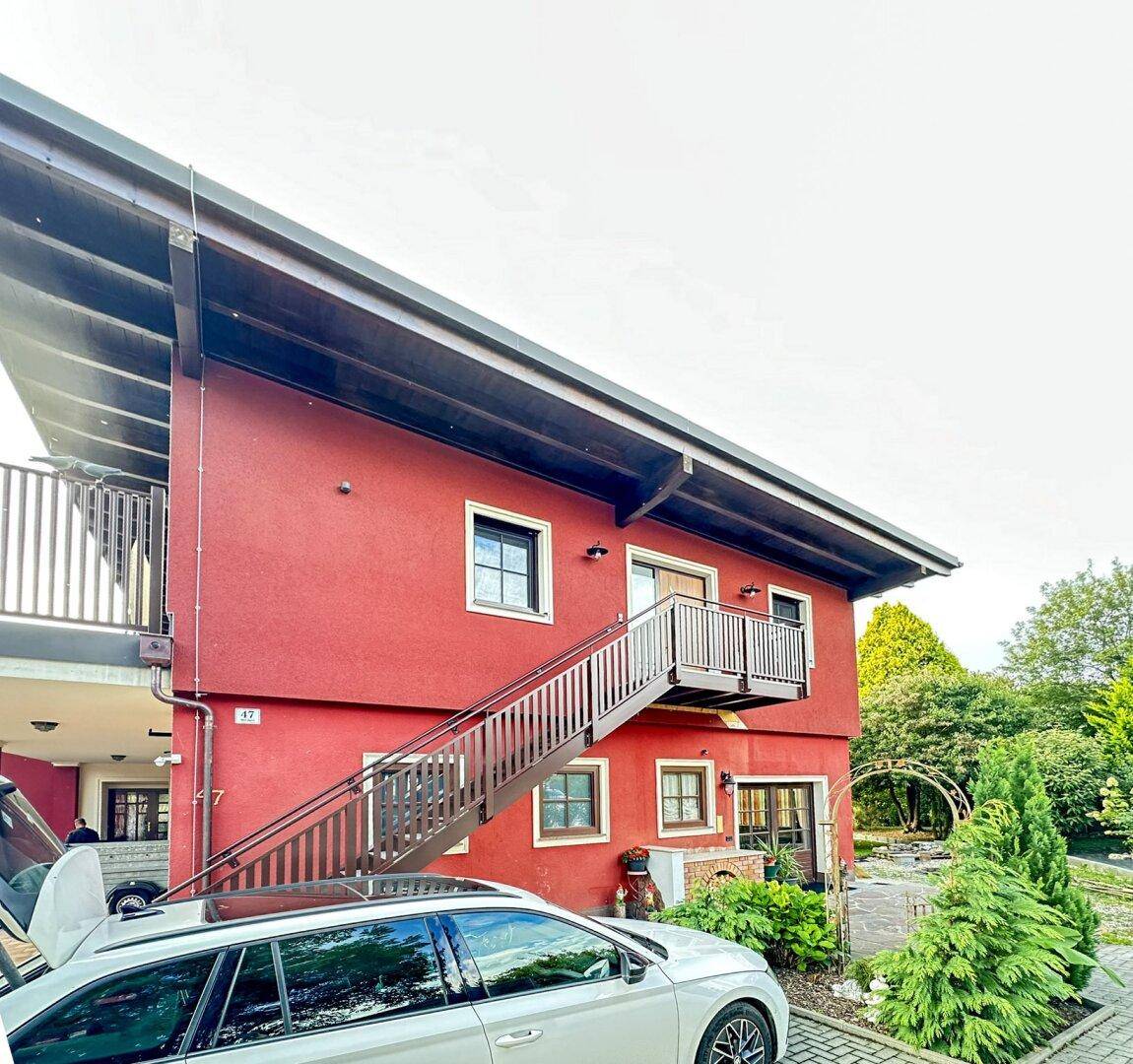 Rotes Gebäude mit Außentreppe, Balkon und gepflegtem Gartenbereich, umgeben von viel Grün.
