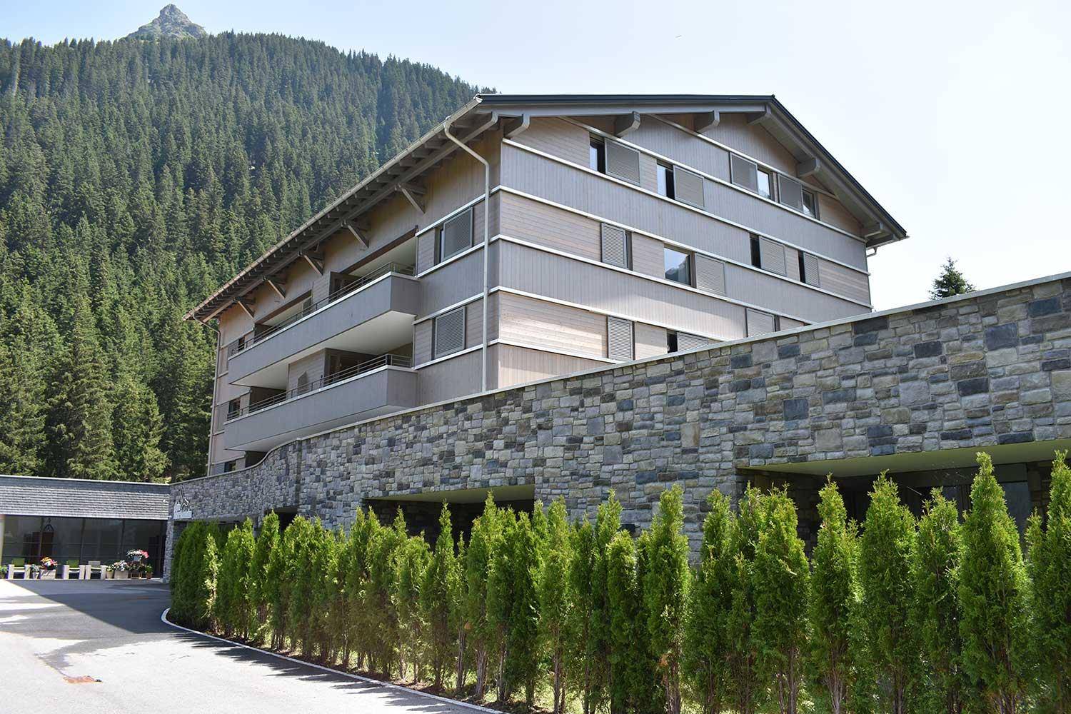 Ansicht des Gebäudes mit Natursteinmauer, gepflegten Hecken und Balkonen in alpiner Umgebung.