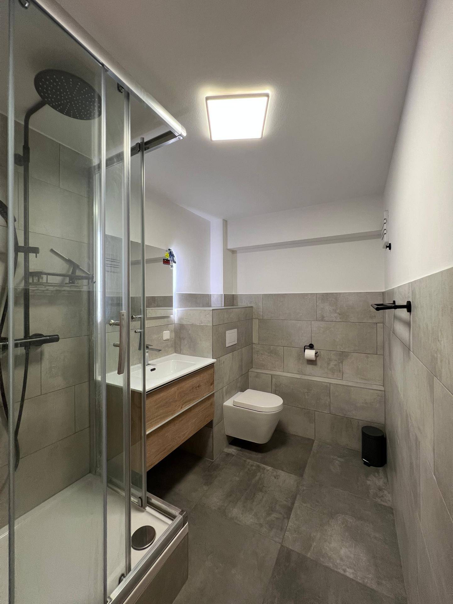 Modernes Badezimmer mit grauen Fliesen, Duschkabine, wandhängendem WC und Holzwaschtischunterschrank.