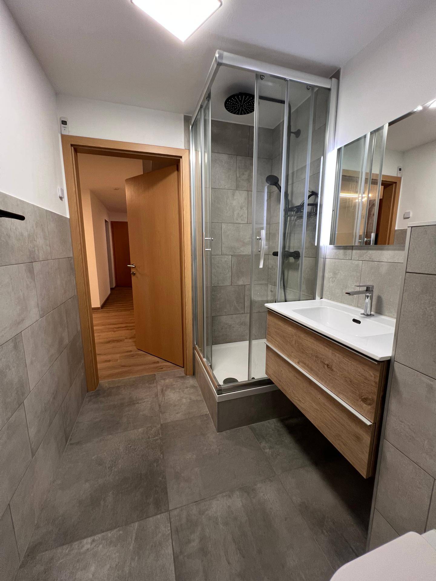 Badezimmer mit Duschkabine, Waschtischunterschrank, beleuchtetem Spiegelschrank und Holztürzugang.