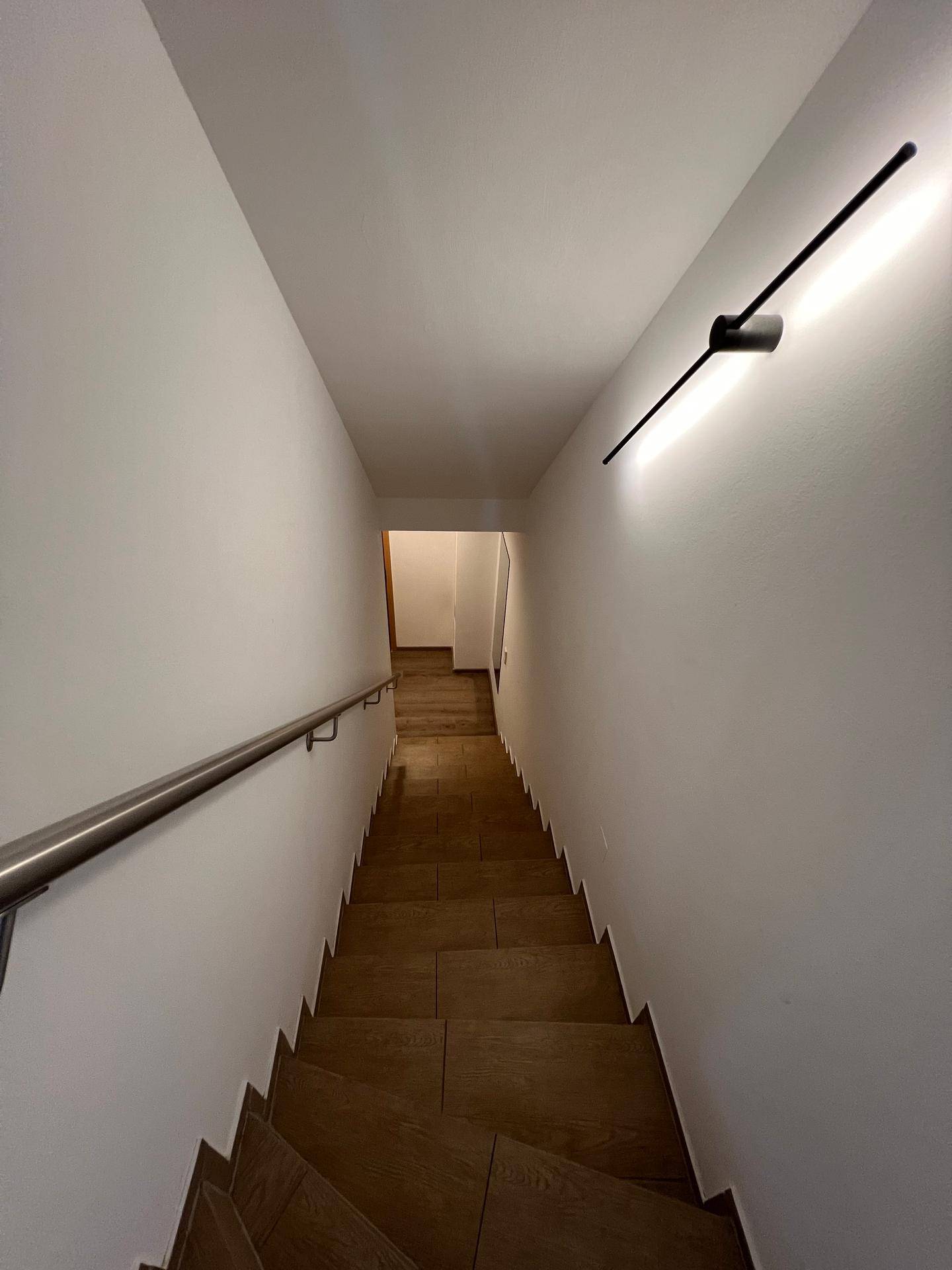Helles Treppenhaus mit Holzstufen, modernem Wandlicht und Edelstahlgeländer führt nach unten.