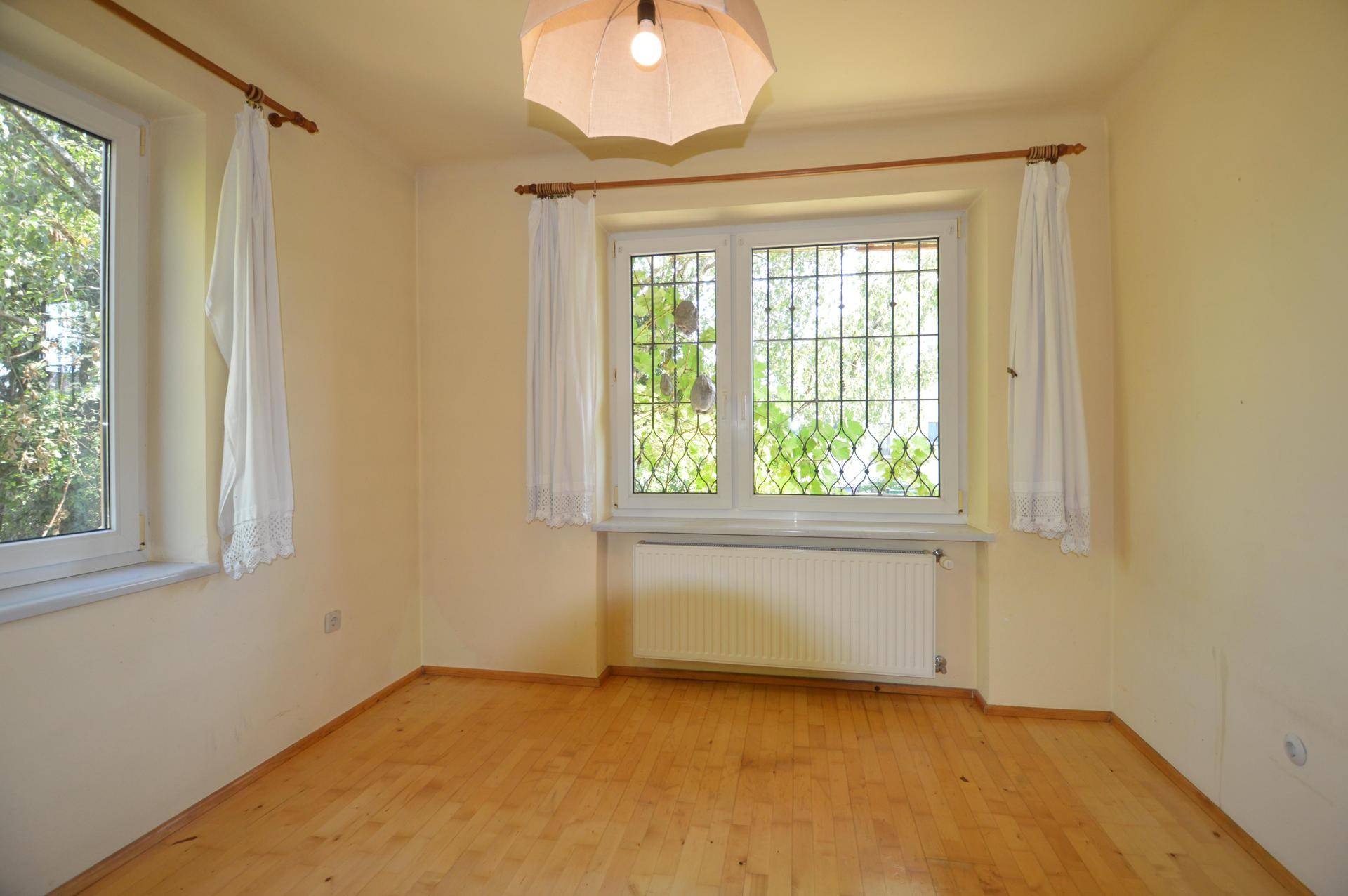 Leeres Zimmer mit hellem Holzboden und einem Fenster mit schmiedeeisernem Gitter und Blick ins Grüne.