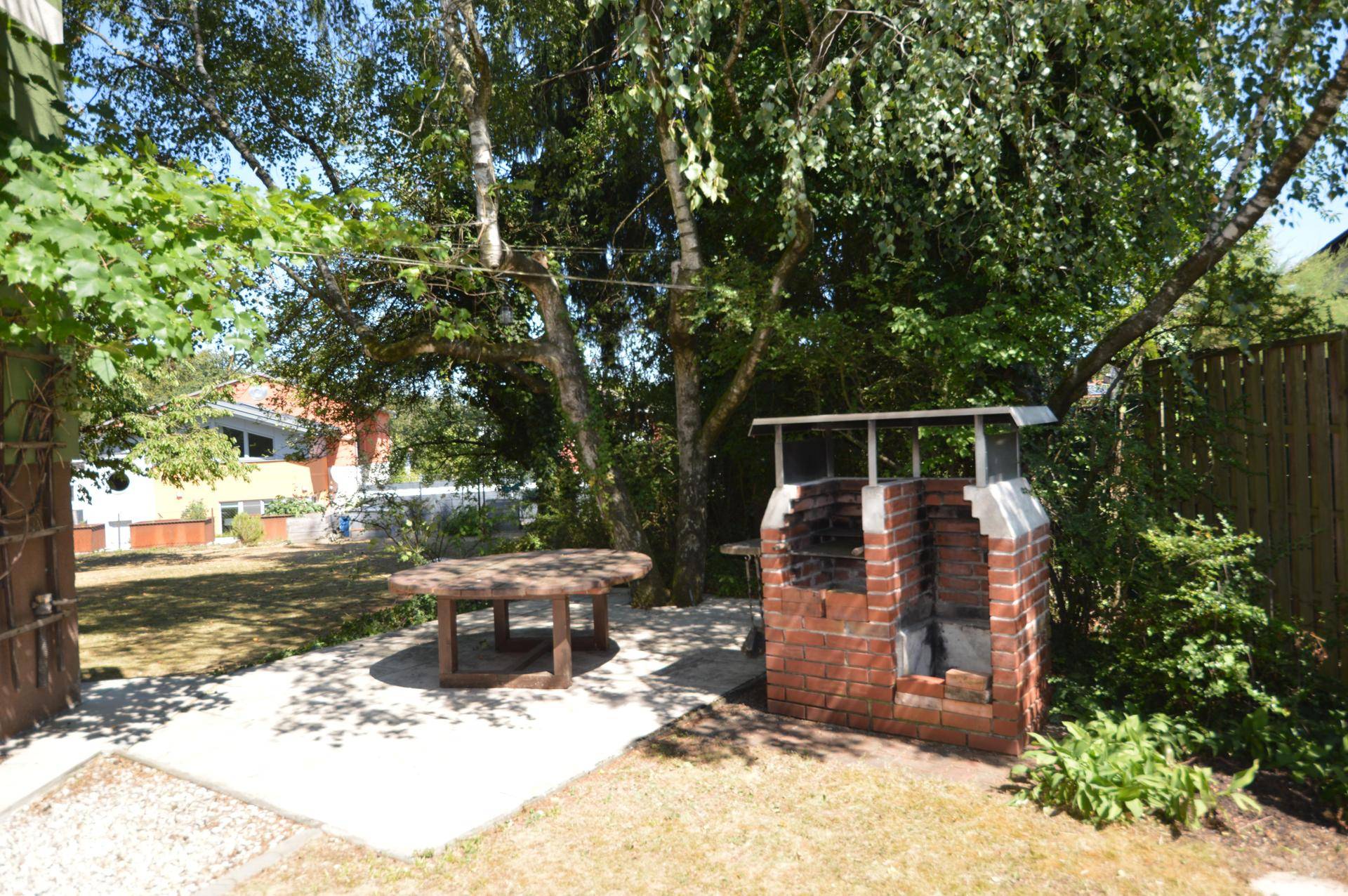 Gemütlicher Grillplatz im Garten mit gemauertem Grill und einem runden Holztisch unter Bäumen.