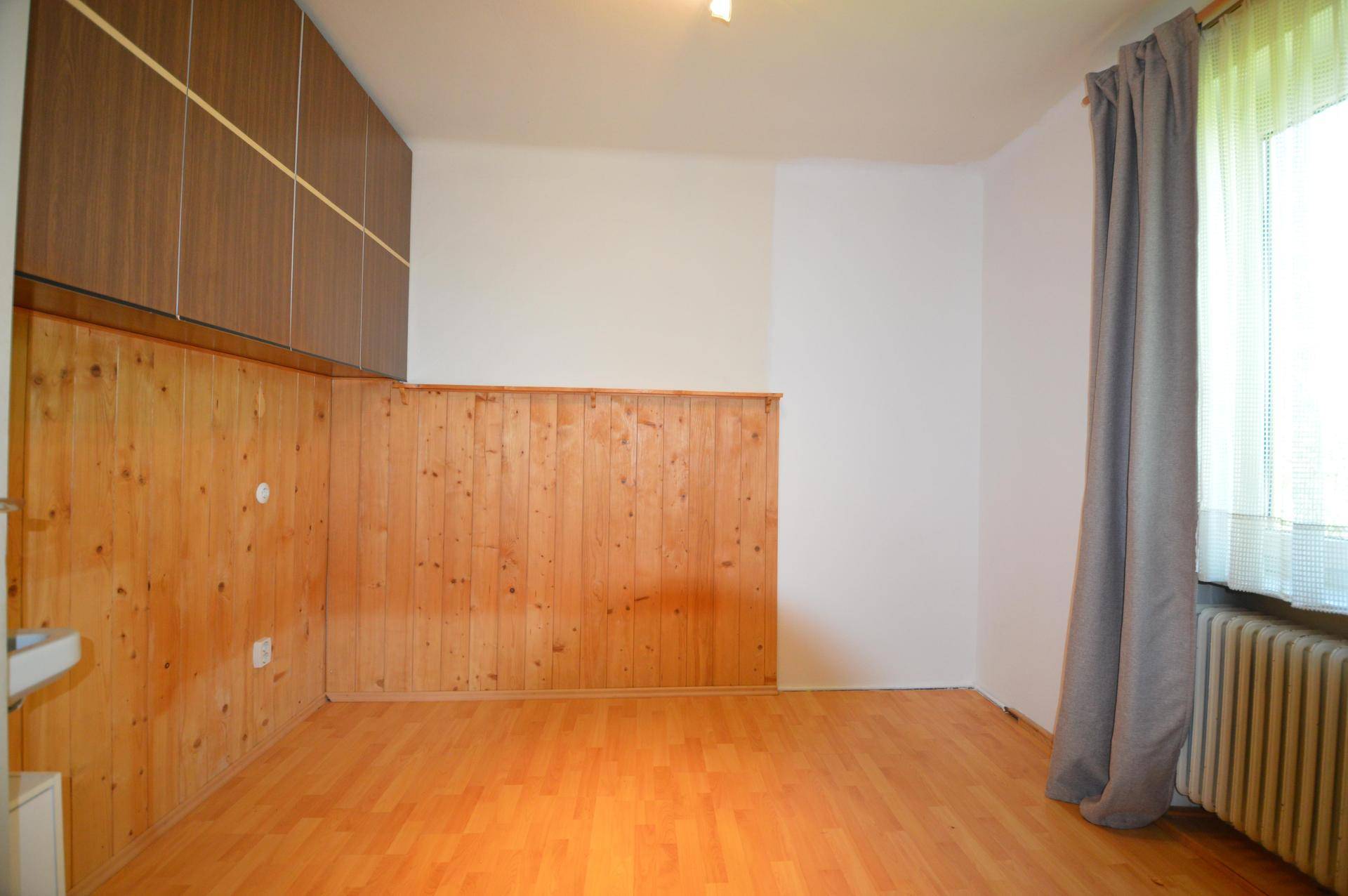 Zimmer mit Holztäfelung an einer Wand, hellem Holzboden und einem Fenster mit Vorhängen.