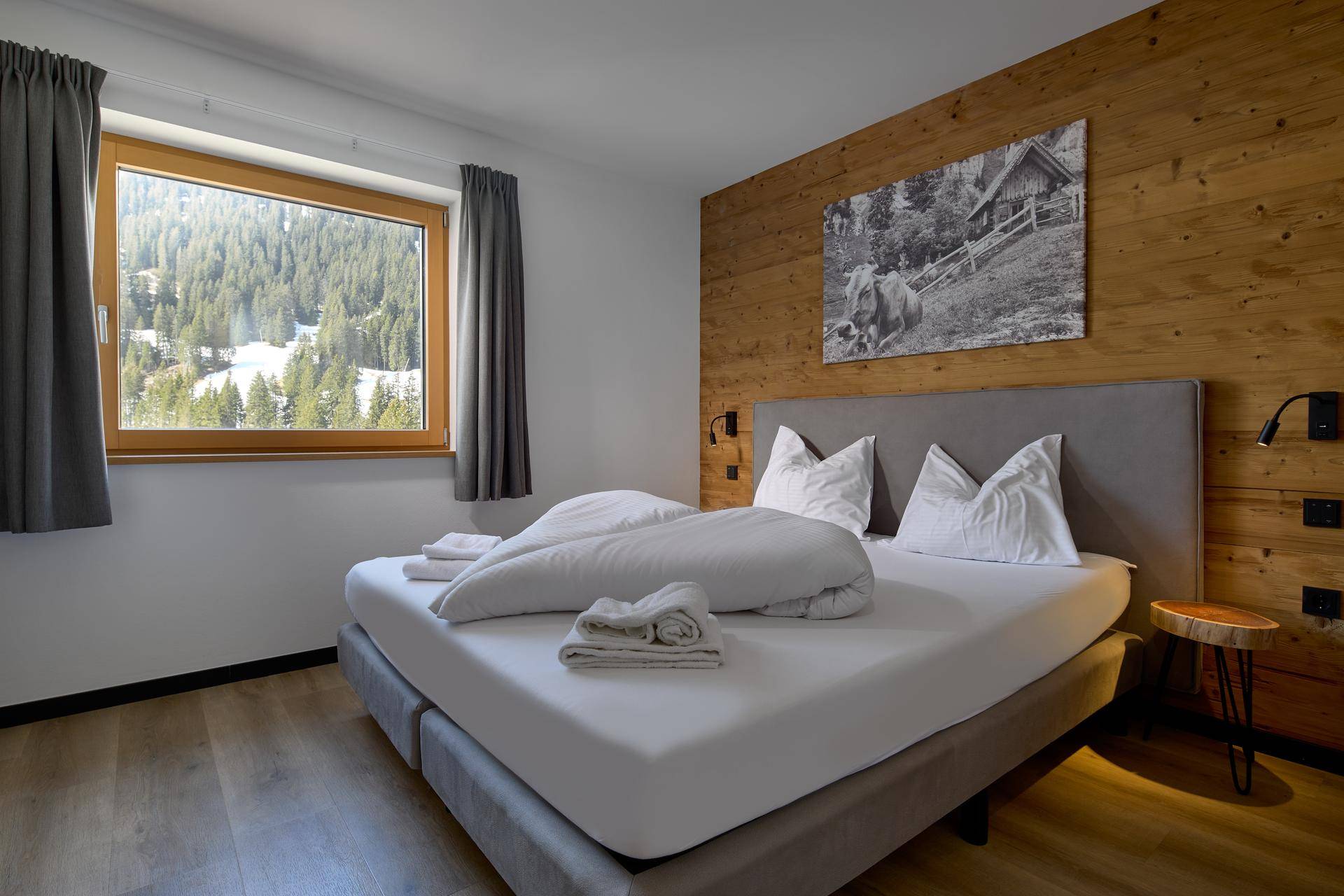 Schlafzimmer mit Holzwand, Doppelbett und Fenster mit Blick auf die verschneite Berglandschaft.