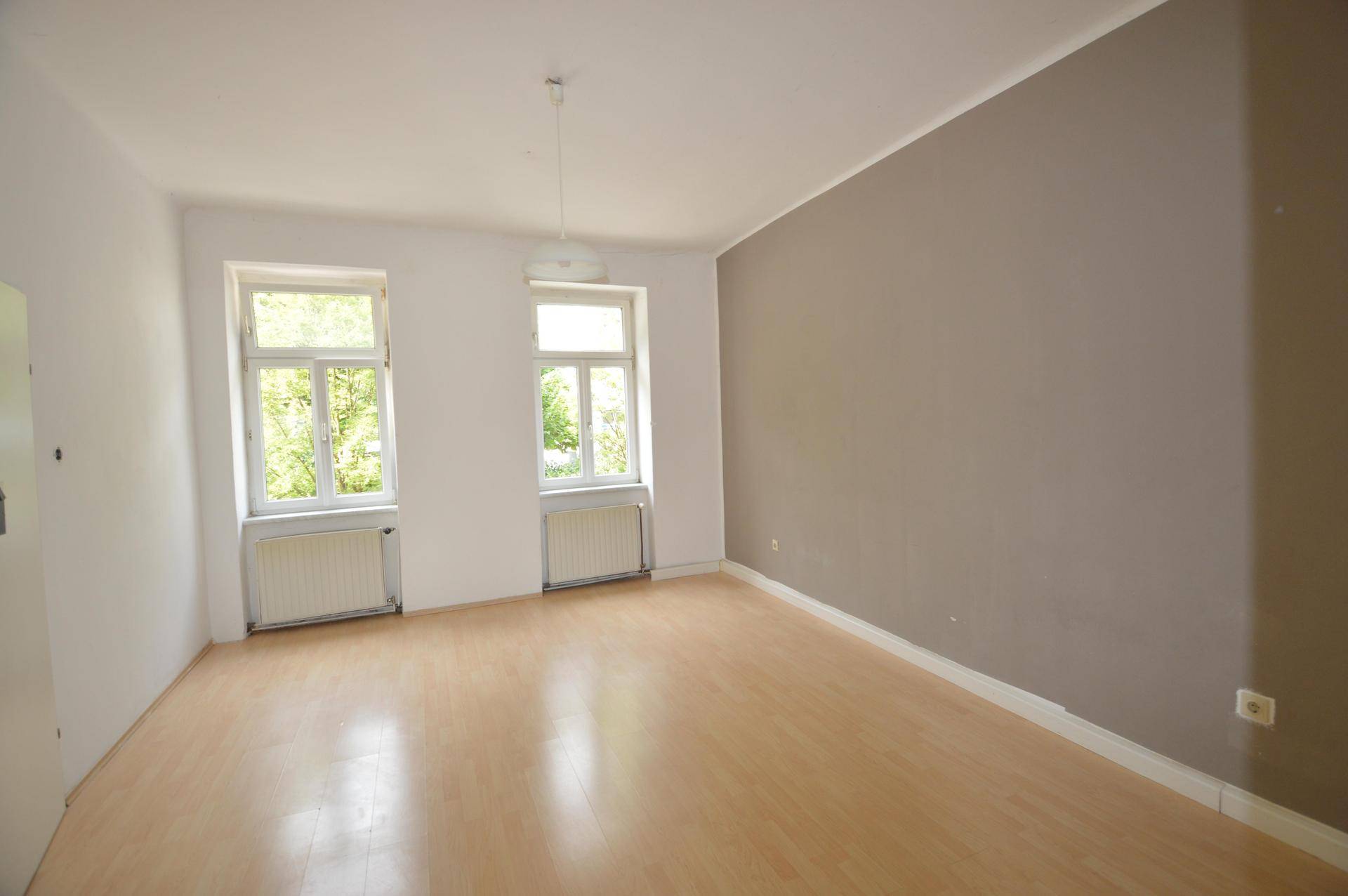 Geräumiges Zimmer mit hellem Laminatboden, einer grauen Akzentwand und zwei Fenstern.