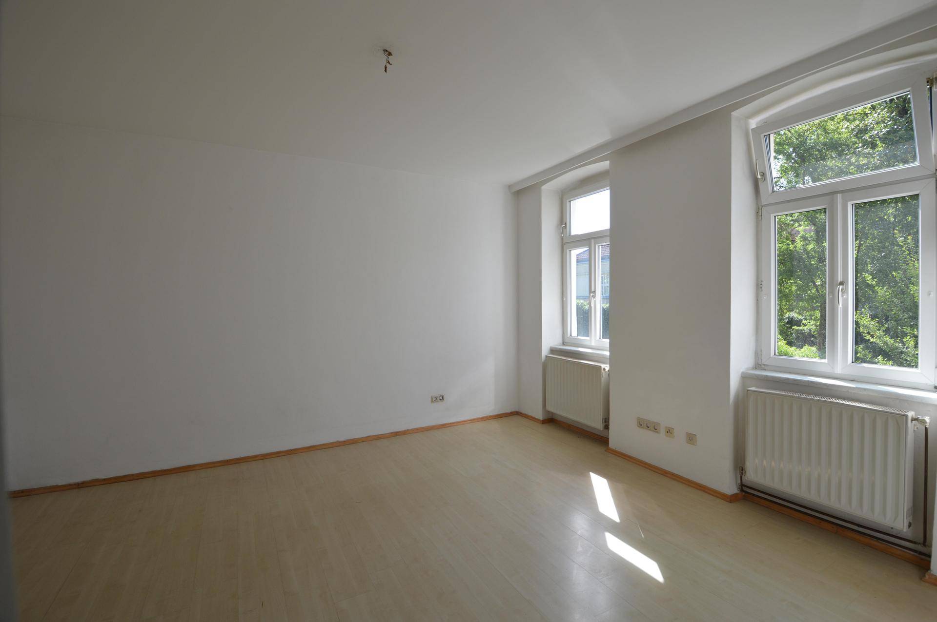 Leeres Zimmer mit hellem Laminatboden, weißen Wänden und zwei Fenstern.