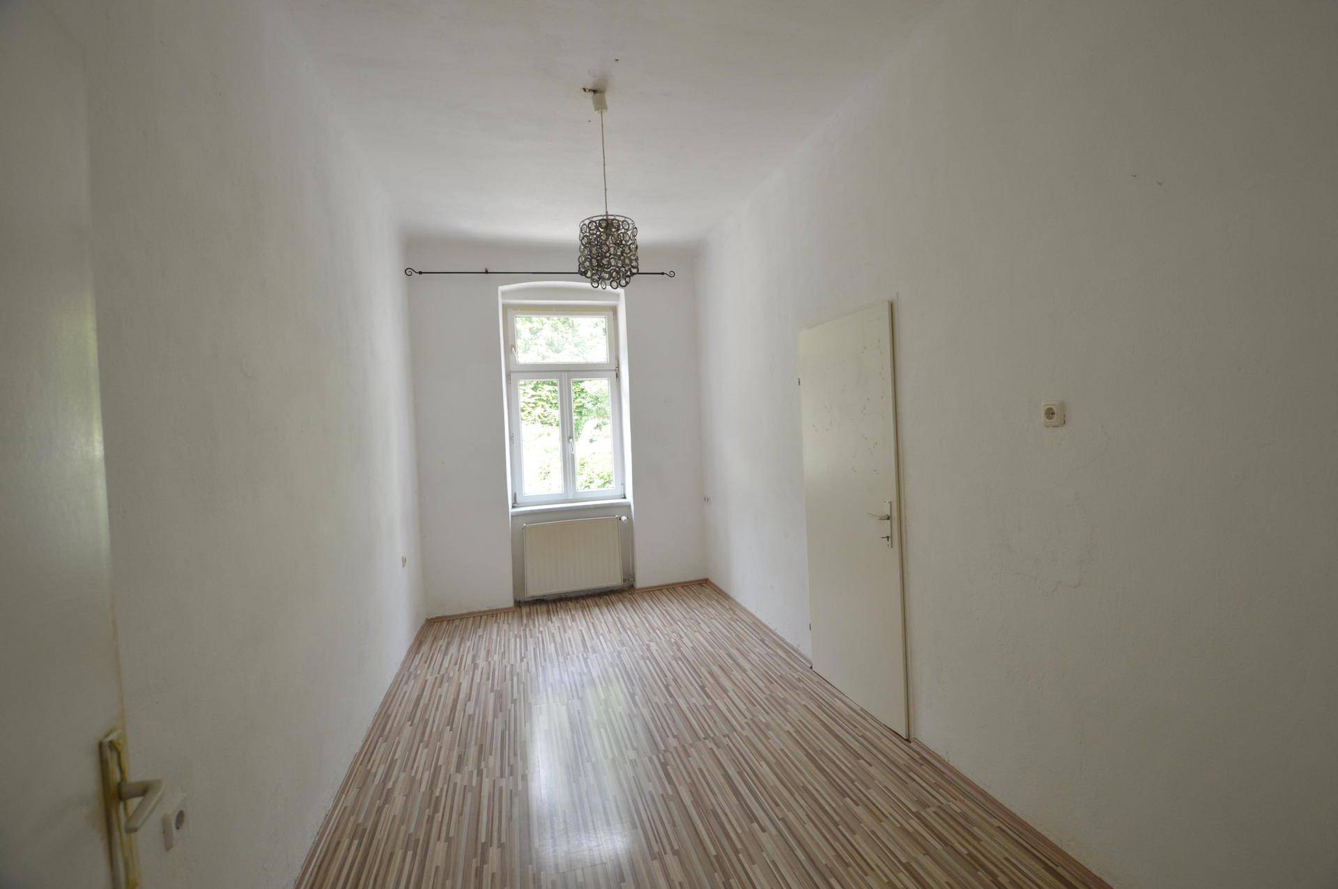 Langes, schmales Zimmer mit Laminatboden, weiß gestrichenen Wänden und einem Fenster.