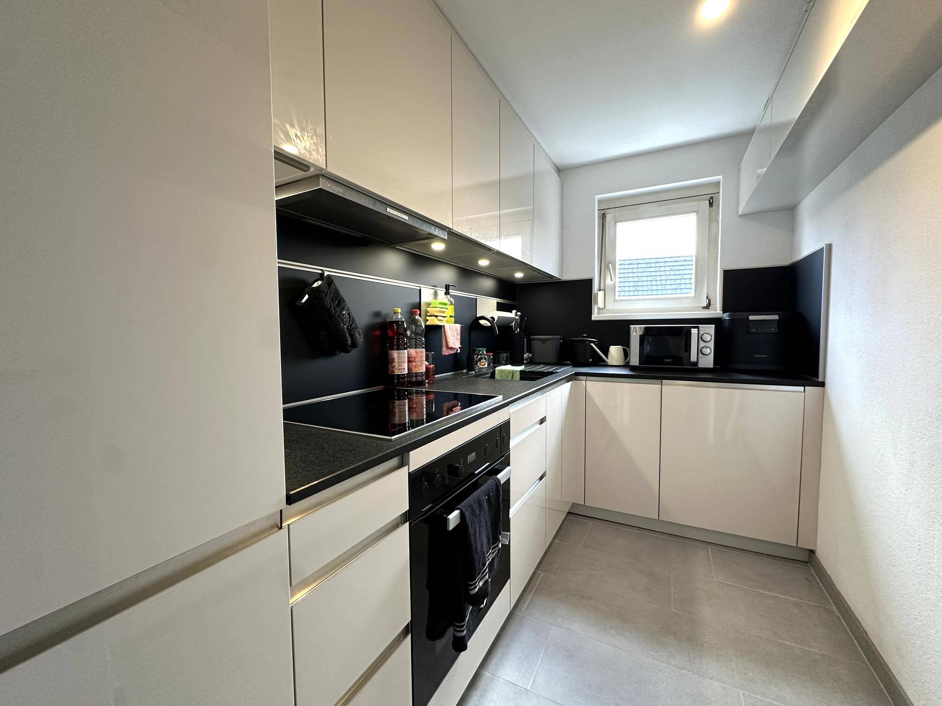 Moderne Küche mit weißen Hochglanzfronten, schwarzen Arbeitsplatten und Fenster.