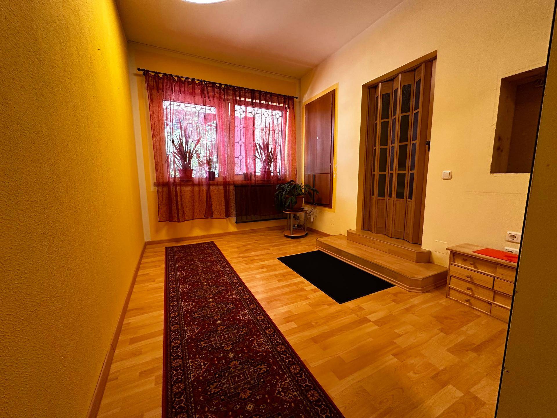 Langer Flur mit Holzboden, gelben Wänden und einem Fenster mit roten Vorhängen.