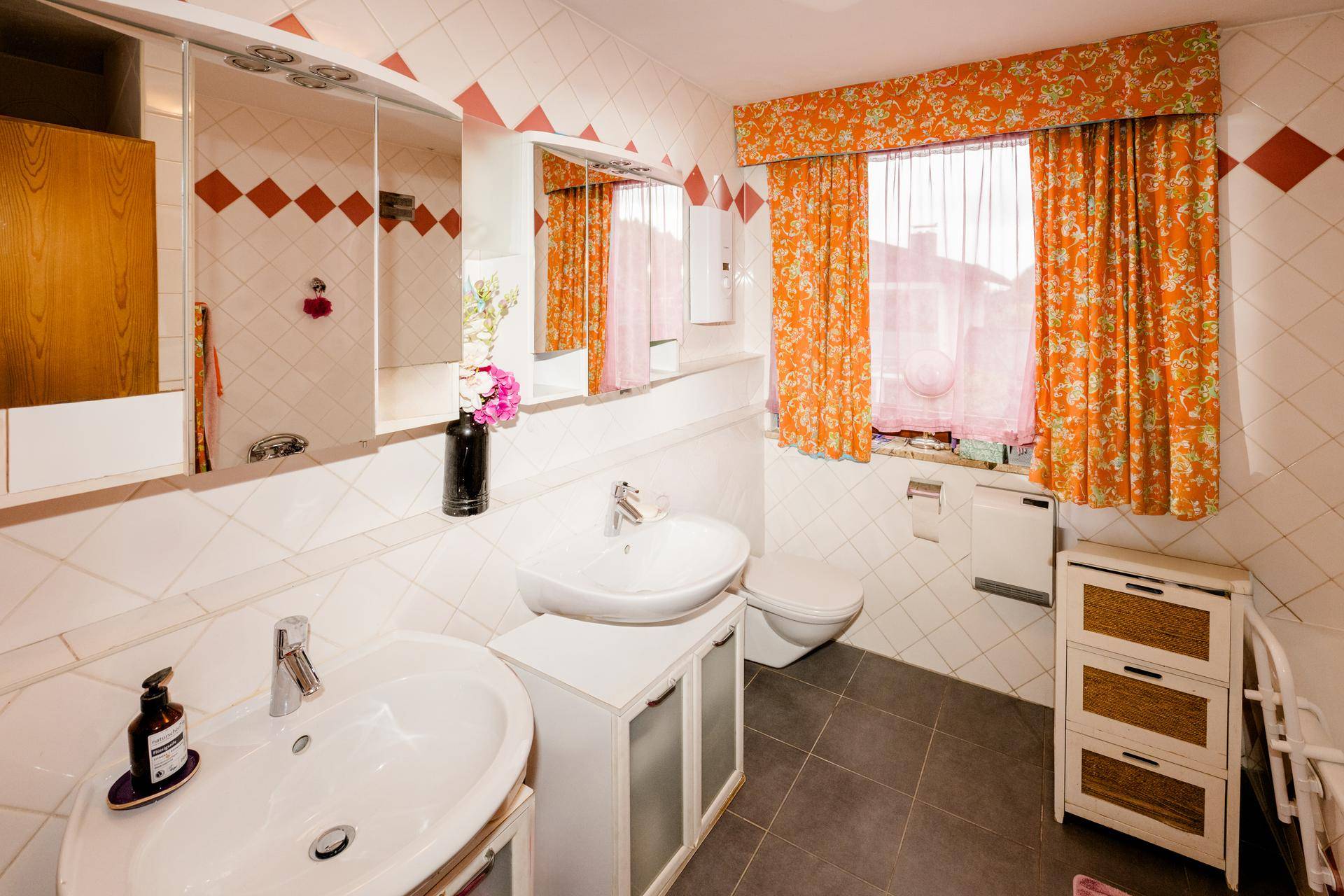 Badezimmer mit zwei Waschbecken, WC, Fenster und weißen Fliesen mit orangefarbenen Akzenten.