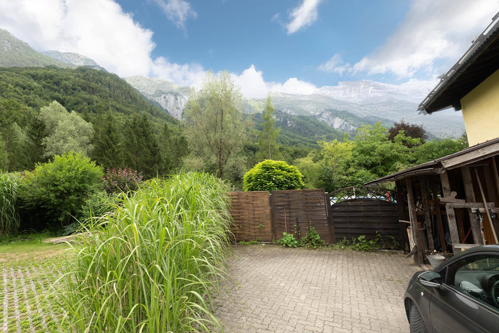 Gepflasterte Auffahrt mit einem Auto, Holzzaun und hohen Gräsern vor einer beeindruckenden Bergkulisse.