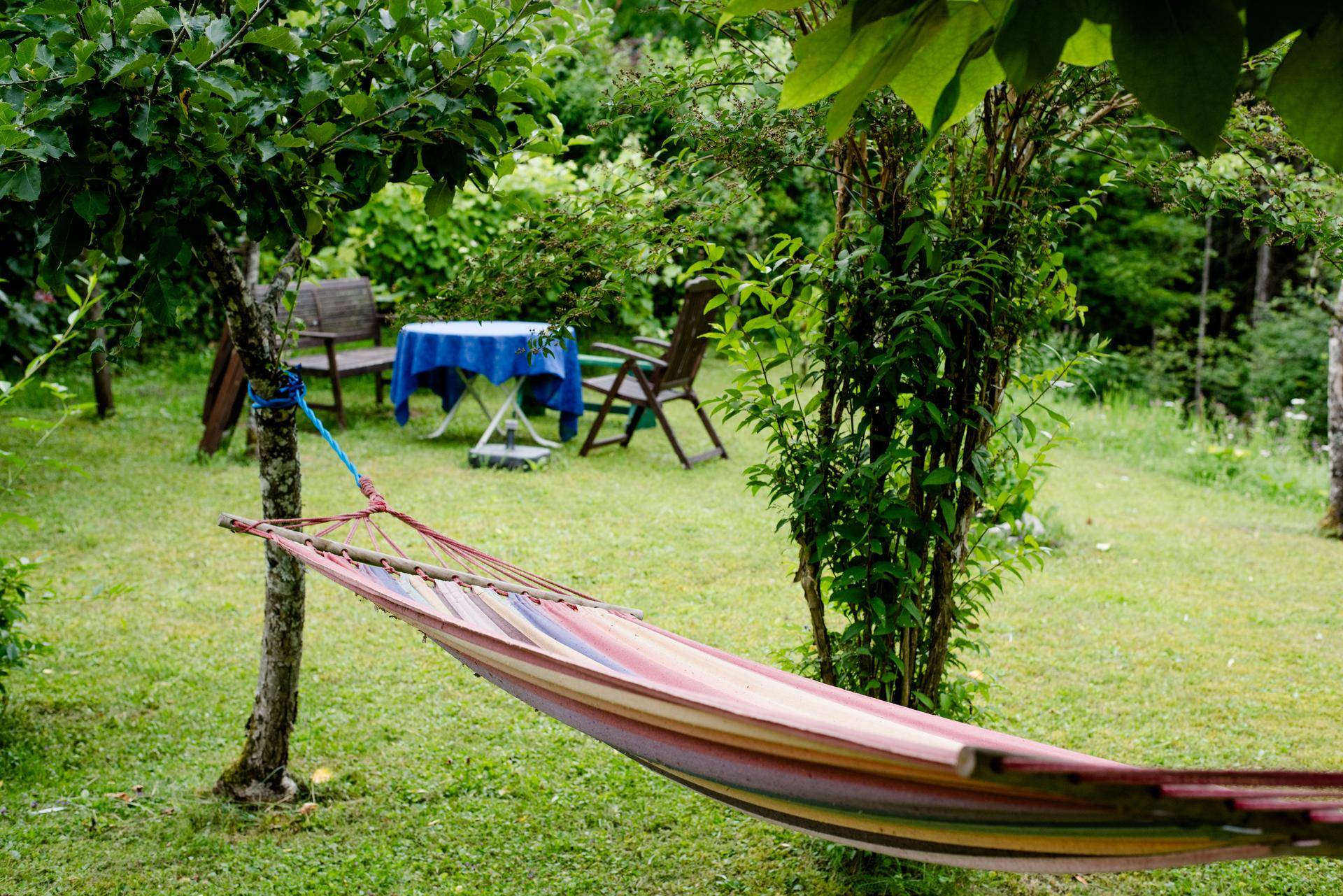 Bunte Hängematte zwischen Bäumen in einem grünen Garten mit einer Sitzecke im Hintergrund.