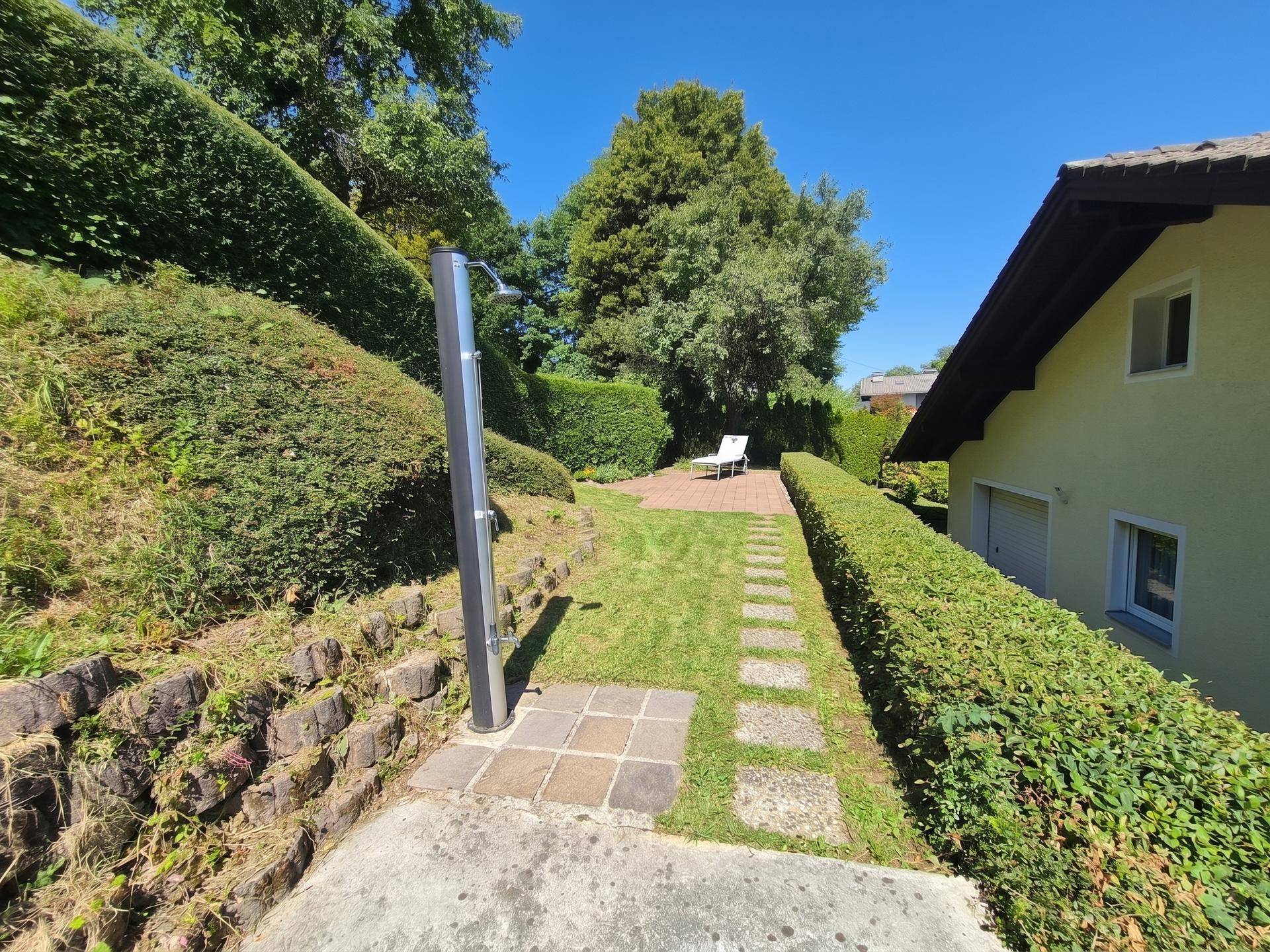 Sonniger Gartenbereich mit Rasenfläche, Steinplattenweg und Außendusche.
