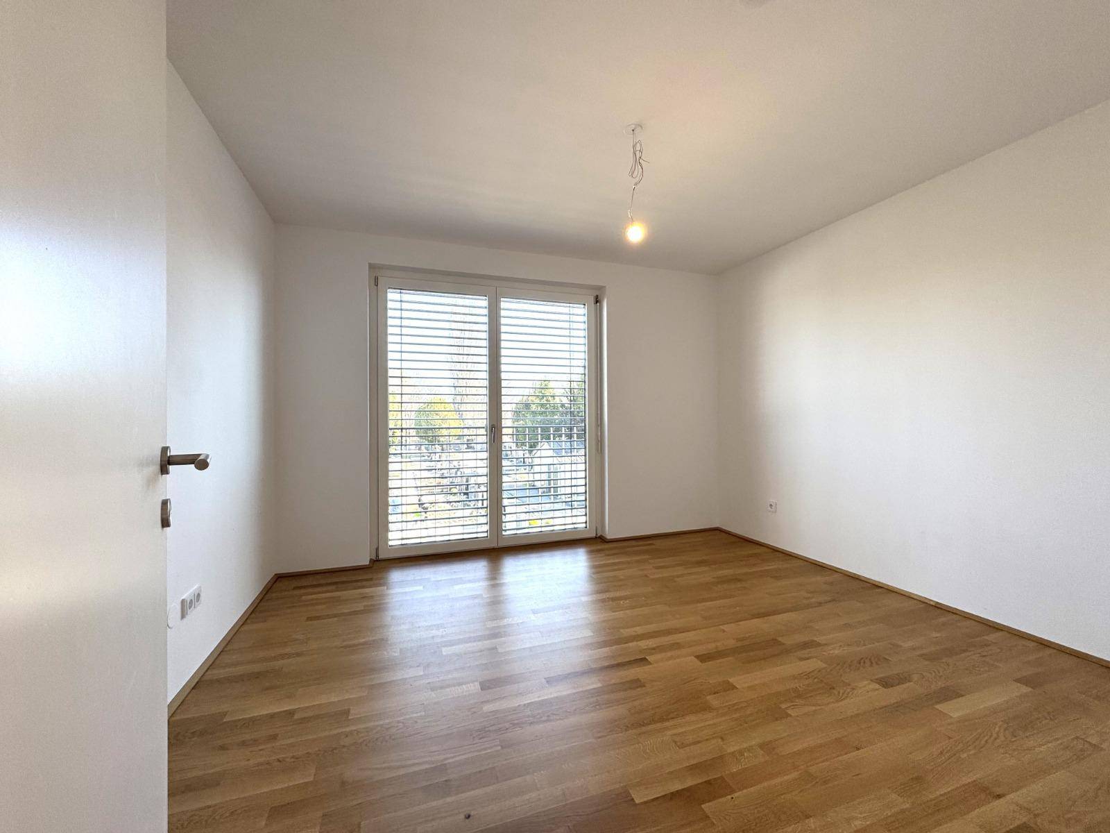 Helles Schlafzimmer mit Holzboden und großem Fenster, das viel Tageslicht hereinlässt.