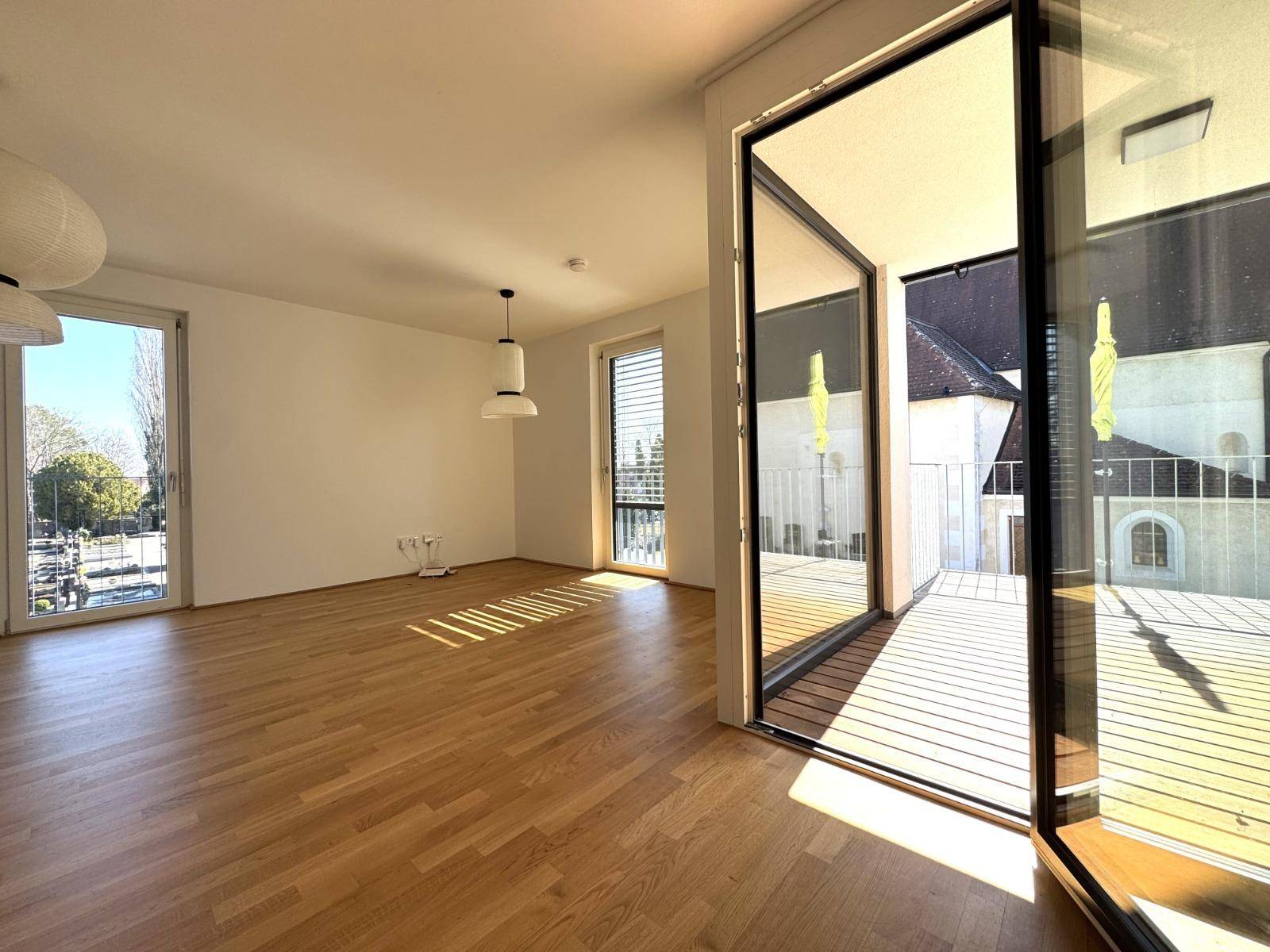 Geräumiges, helles Wohnzimmer mit Holzboden und großen Fenstern, die auf einen Balkon führen.