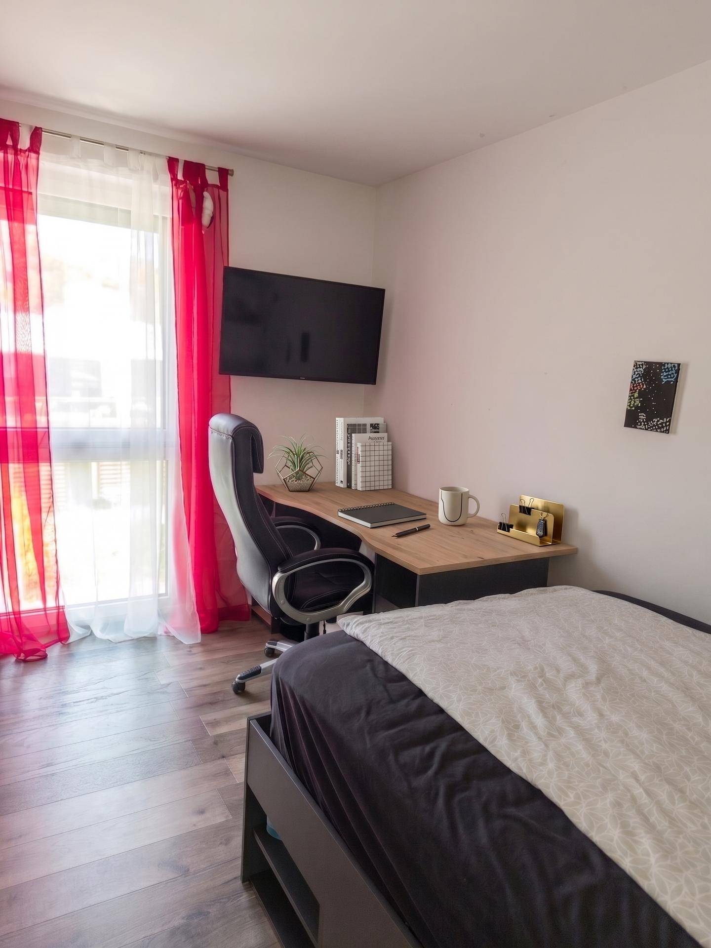 Helles Schlafzimmer mit Schreibtisch, Fernseher und Zugang zum Balkon.