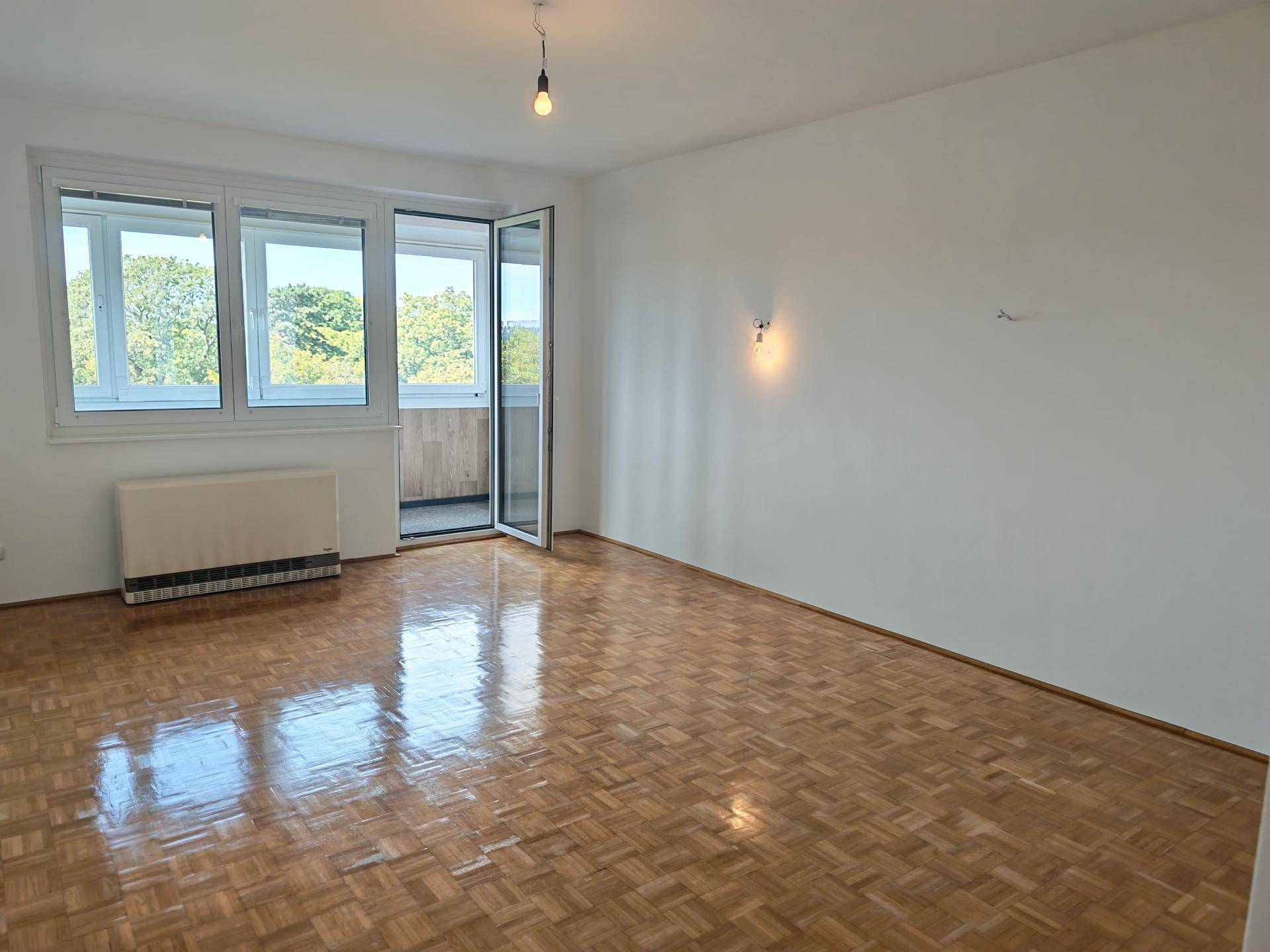 Helles, leeres Wohnzimmer mit glänzendem Parkettboden und Zugang zu einem kleinen Balkon.