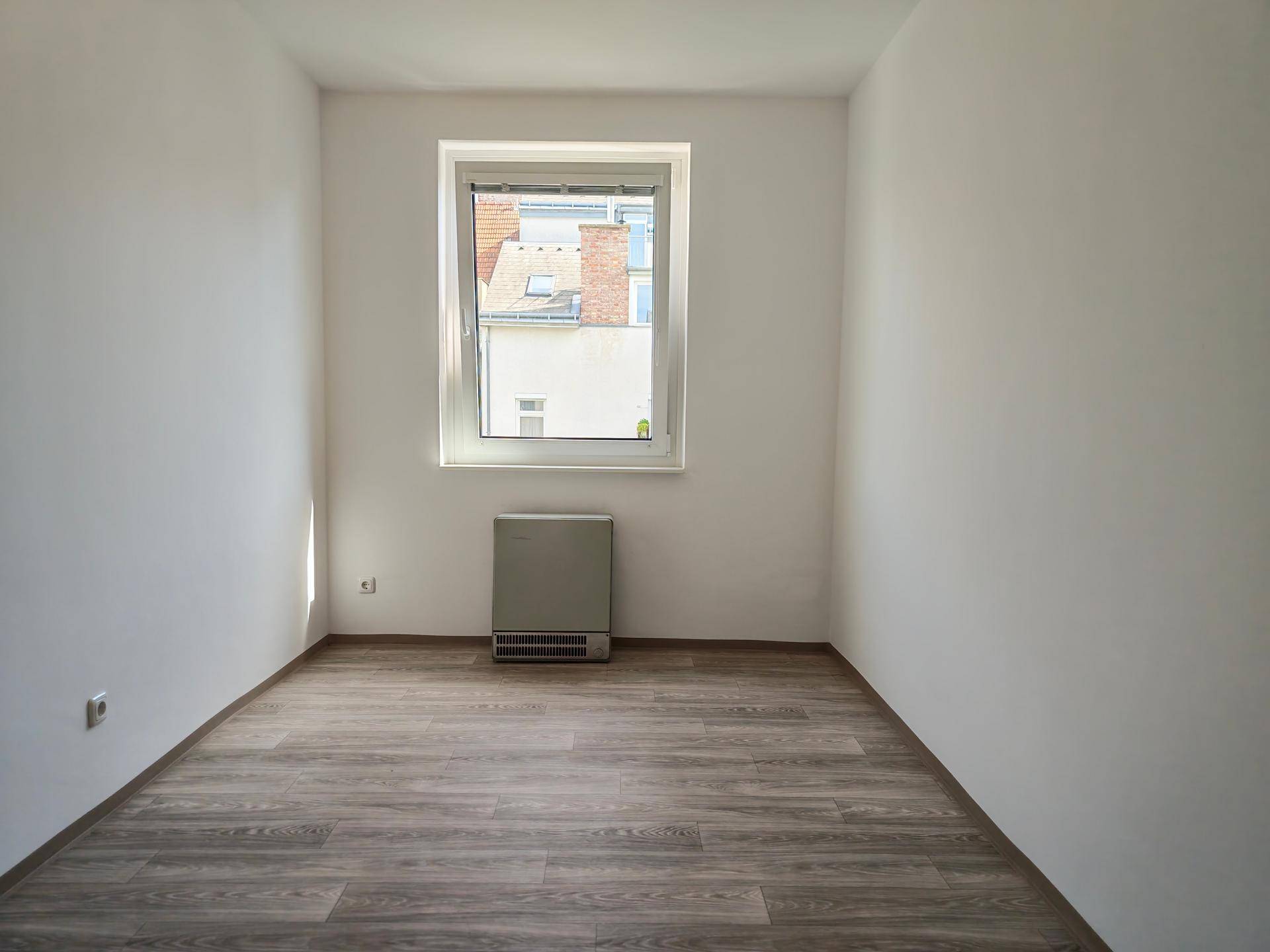 Kleines, leeres Zimmer mit Laminatboden und einem Fenster, das Tageslicht hereinlässt.