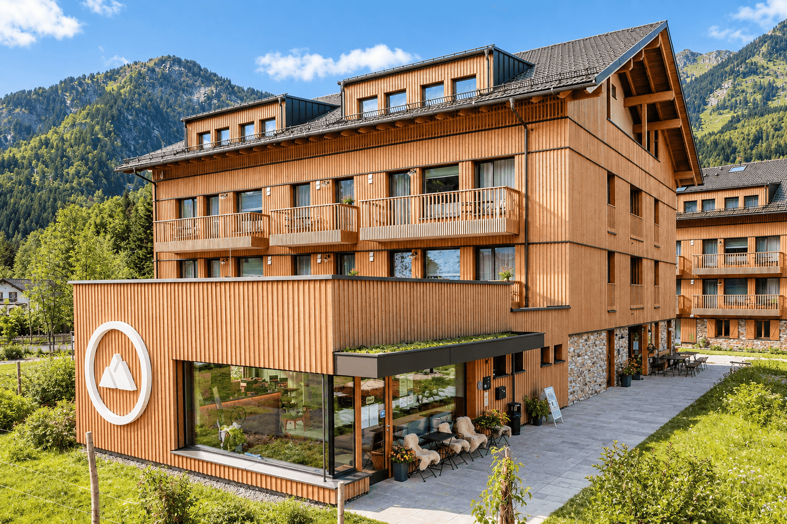 Modernes Aparthotel mit Holzfassade und Außenterrasse vor alpiner Bergkulisse.