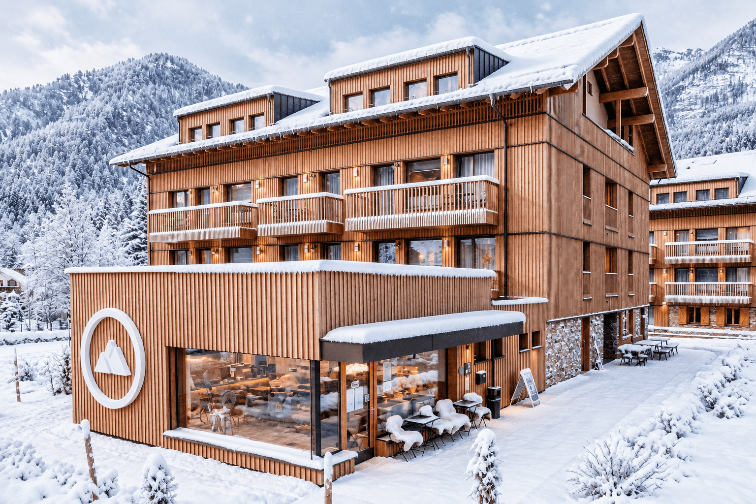 Modernes Holzgebäude des Aparthotels mit Balkonen und großen Fenstern, umgeben von Schnee und Bergen.