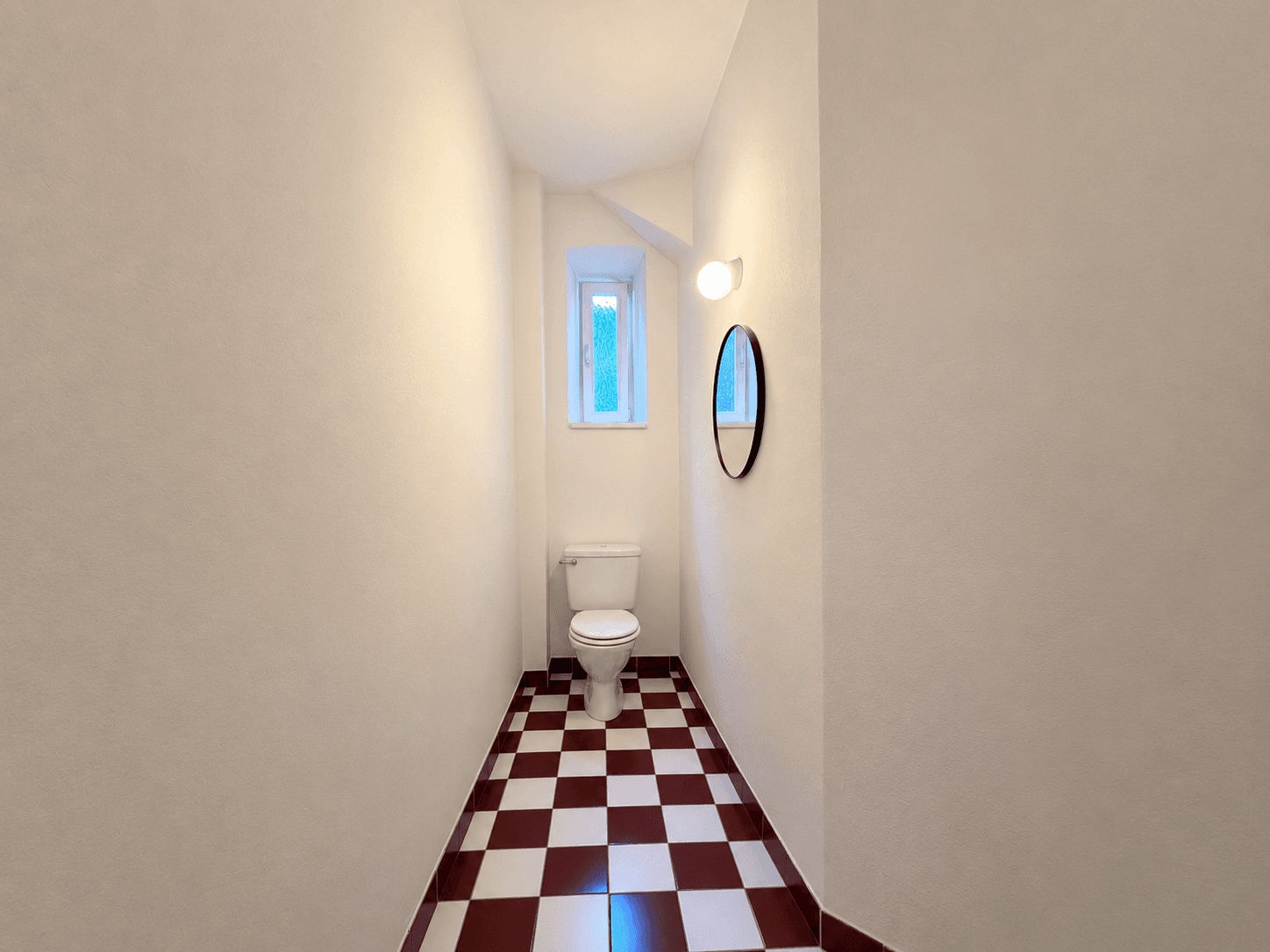 Schmale Toilette mit rot-weiß karierten Fliesen und einem kleinen Fenster für Tageslicht.