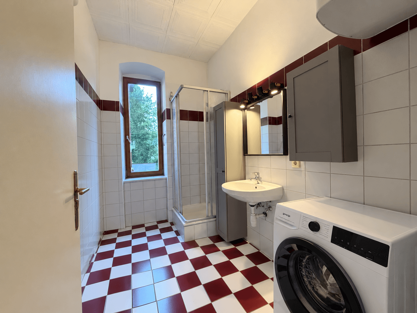 Badezimmer mit rot-weiß karierten Fliesen, Dusche und Waschmaschine, hell durch ein Fenster.
