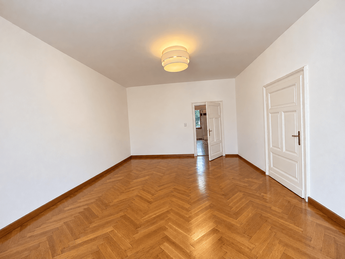 Geräumiges, leeres Zimmer mit Fischgrätparkett und weiß gestrichenen Wänden, bereit zur Einrichtung.