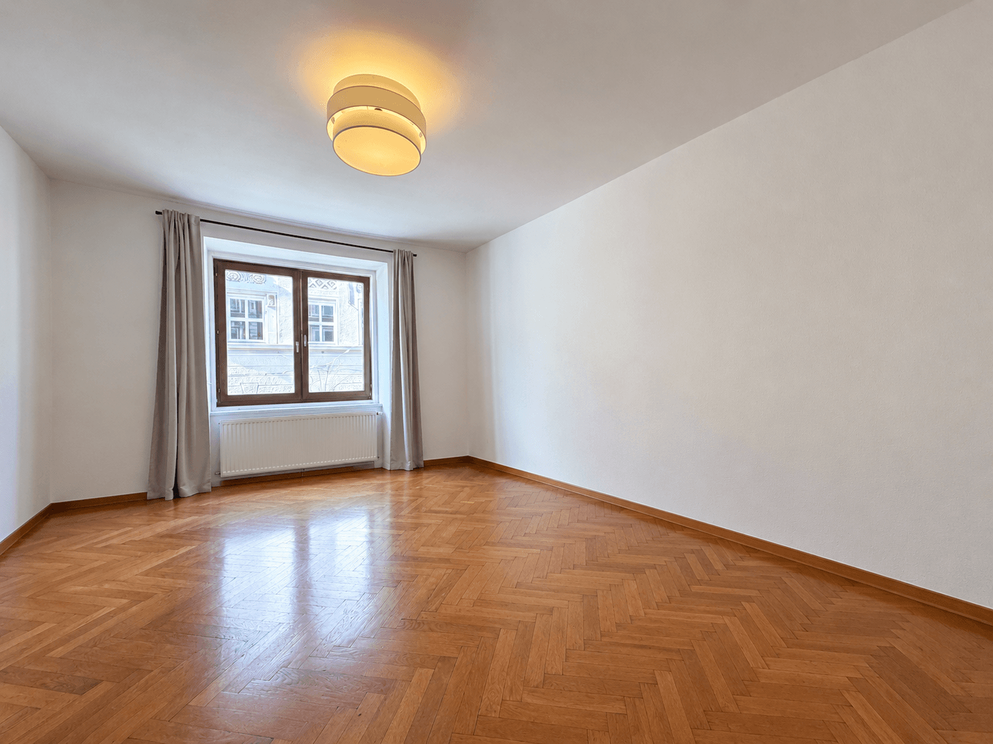 Helles Zimmer mit klassischem Fischgrätparkett und einem Fenster mit Blick auf die Stadt.