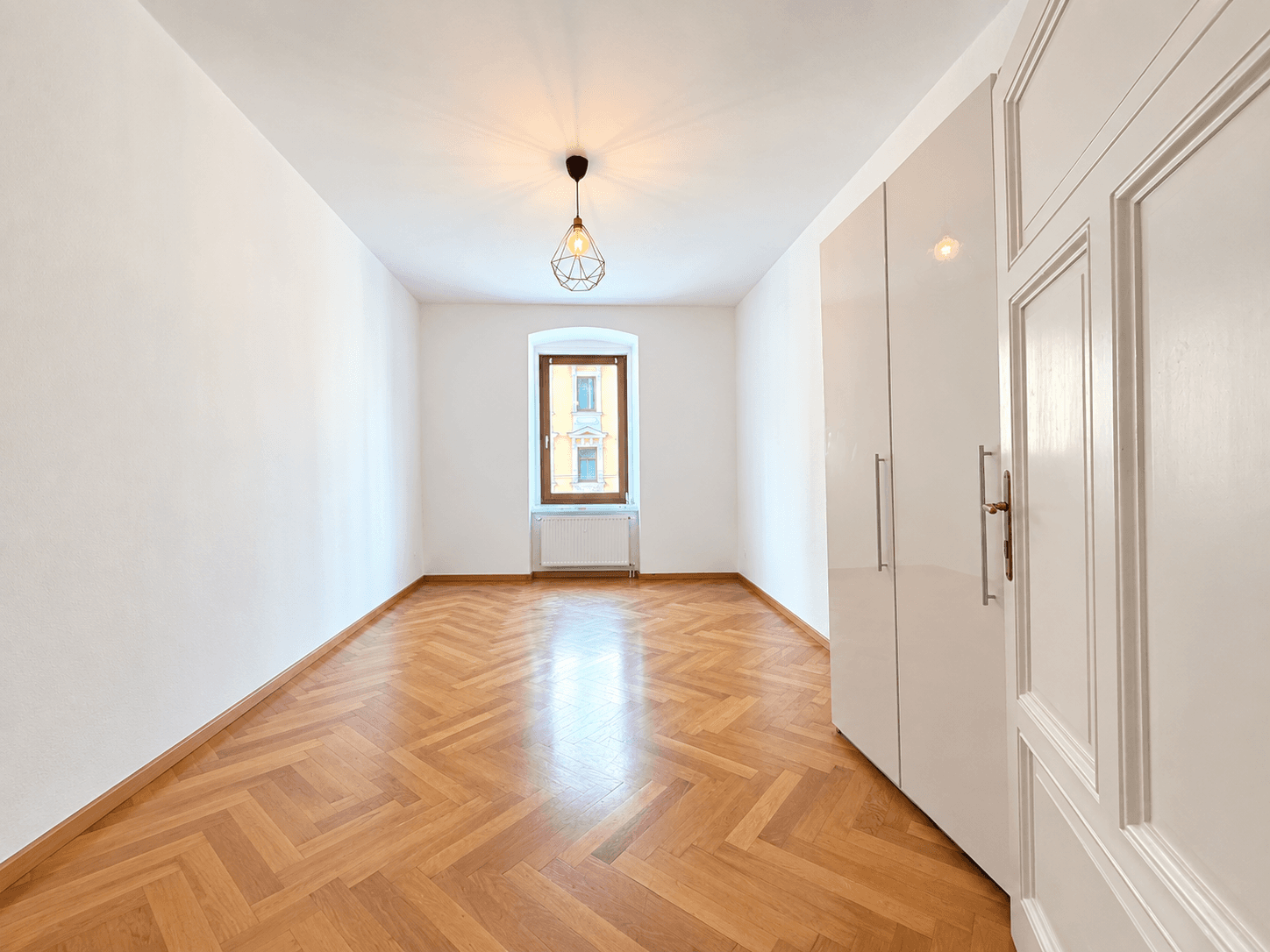 Kleineres Zimmer mit Fischgrätparkett, Einbauschrank und einem Fenster, das viel Licht hereinlässt.
