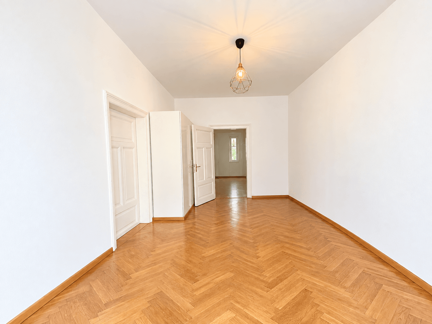 Leeres Zimmer mit Fischgrätparkett und mehreren Türen, die zu anderen Bereichen der Wohnung führen.