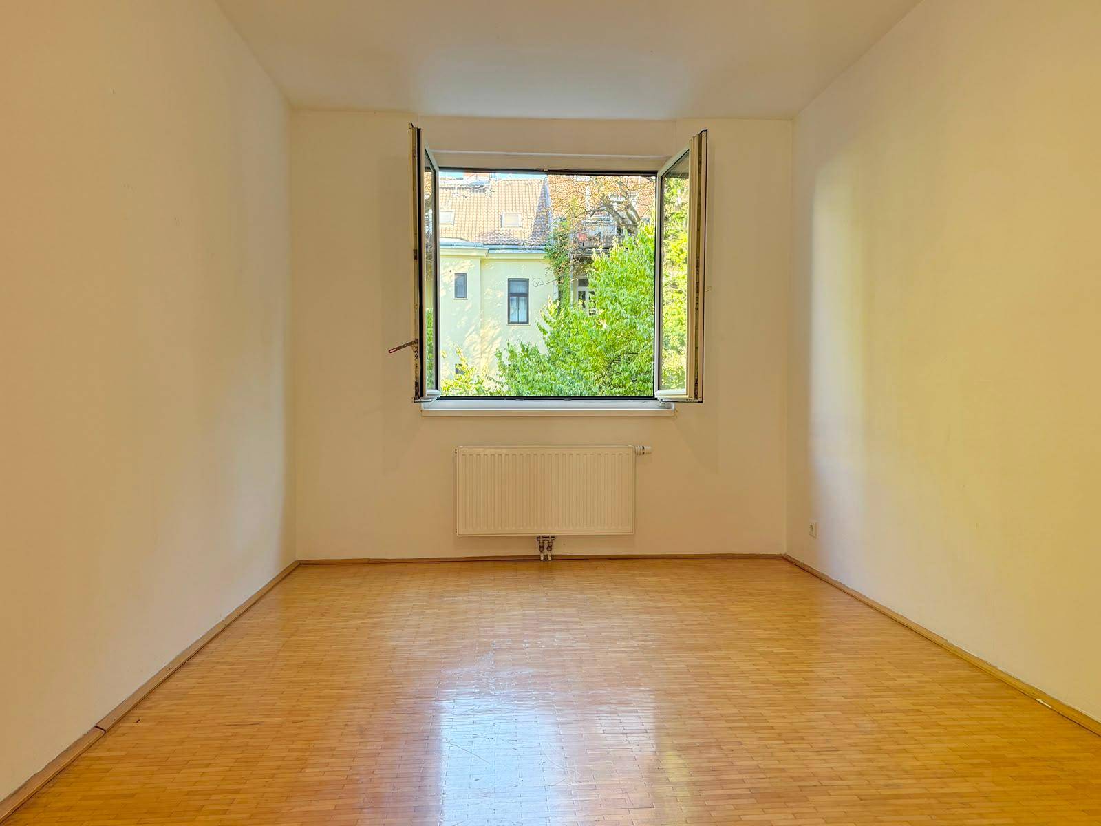 Helles Schlafzimmer mit Parkettboden, großem Fenster und Blick ins Grüne.