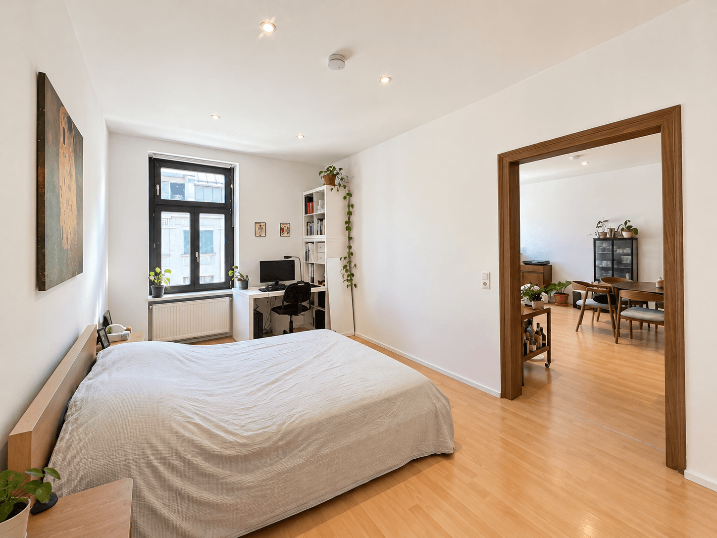 Schlafzimmer mit Holzbett und Schreibtisch, mit Blick durch eine offene Tür in einen Essbereich.