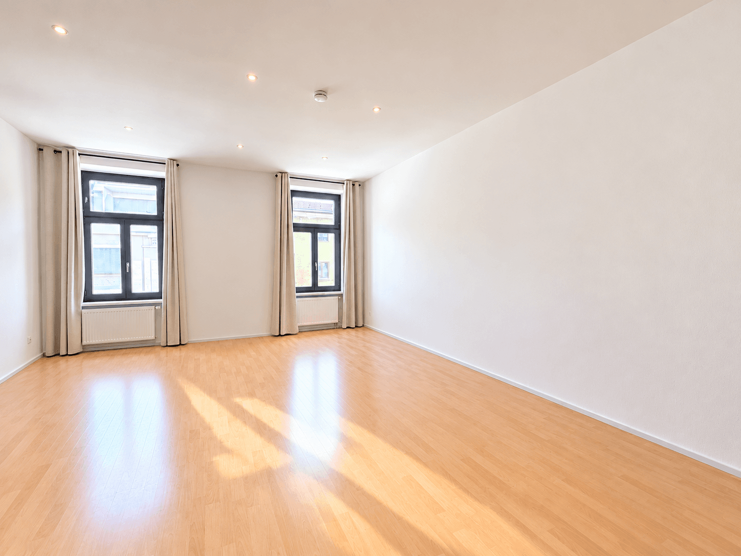 Geräumiges, unmöbliertes Wohnzimmer mit hellem Parkettboden und Einbaustrahlern an der Decke.