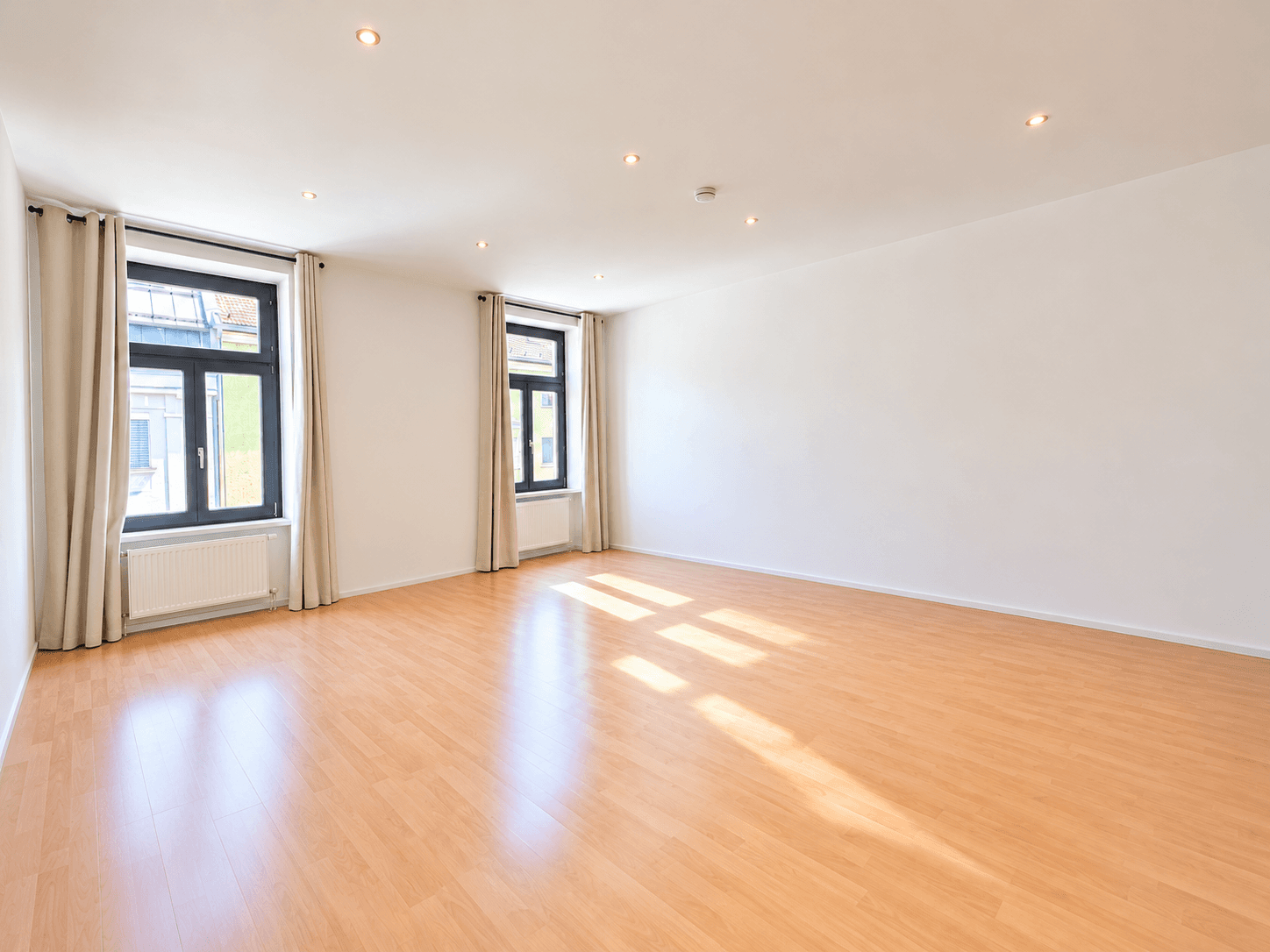 Helles, leeres Wohnzimmer mit Parkettboden und zwei Fenstern, die viel Tageslicht hereinlassen.