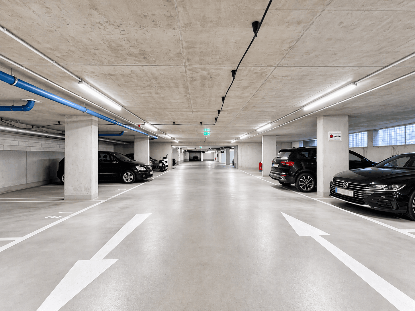 Geräumige und helle Tiefgarage mit Betonwänden und -decken sowie modernen Leuchten und geparkten Autos.
