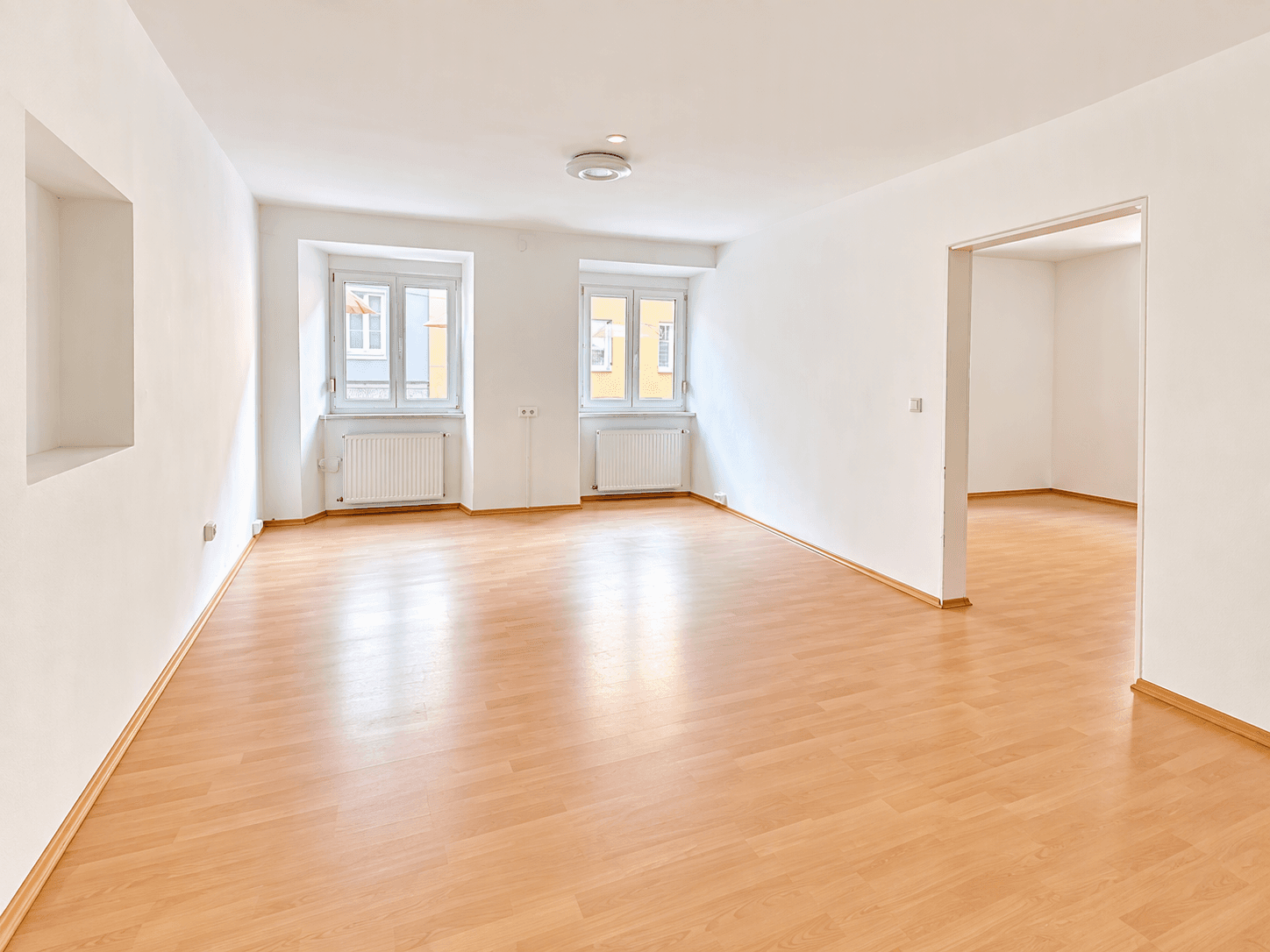 Geräumiges, leeres Zimmer mit hellem Laminatboden und zwei Fenstern.