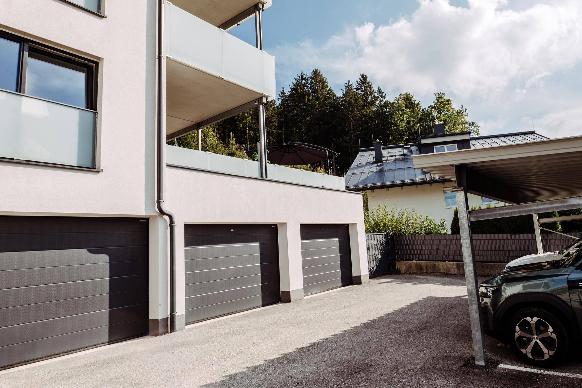 Gebäudefassade mit Garagen und Carport, umgeben von Bäumen und Grünflächen.