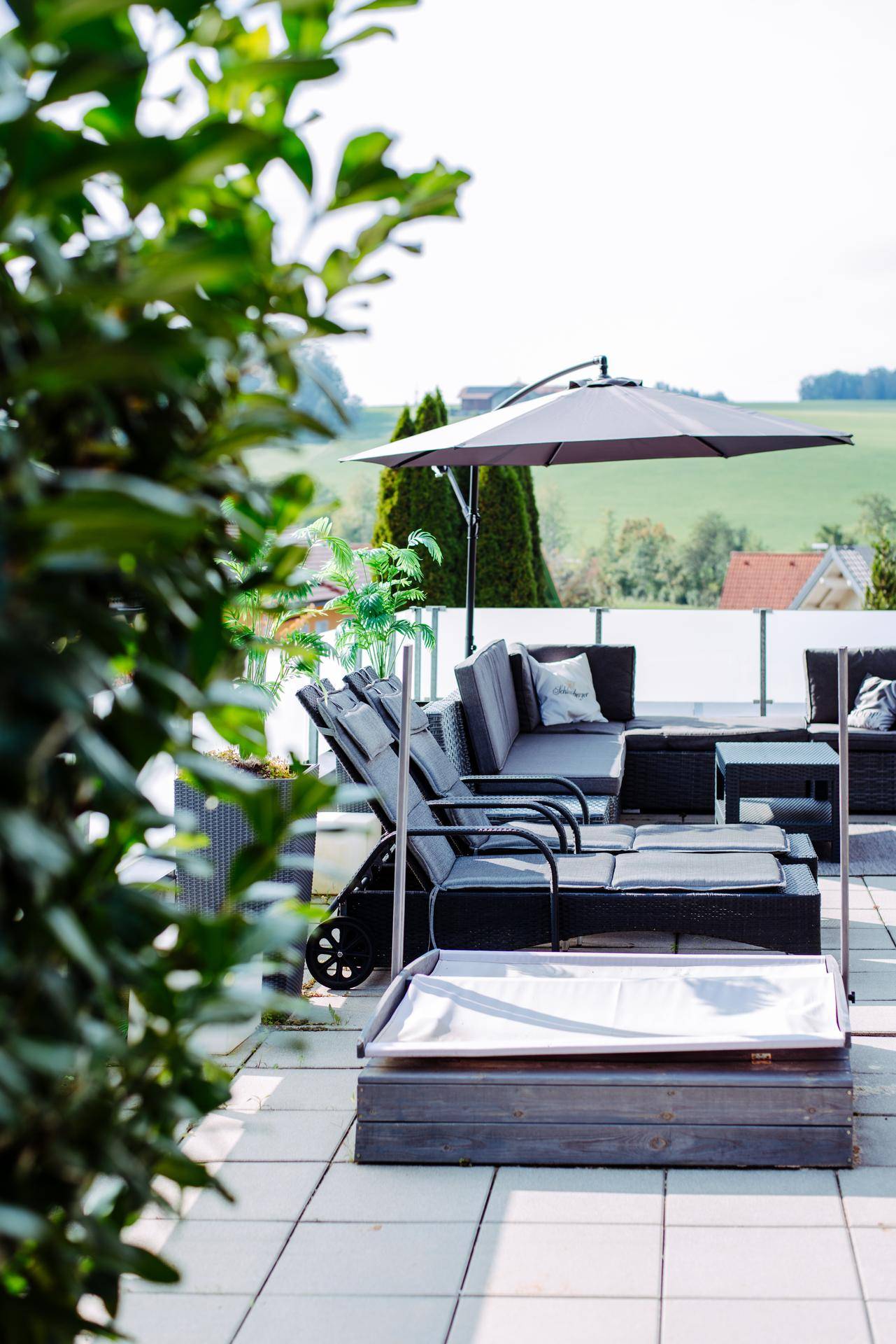 Helle Terrasse mit Loungemöbeln und Sonnenschirm, umgeben von grüner Bepflanzung und Weitblick.
