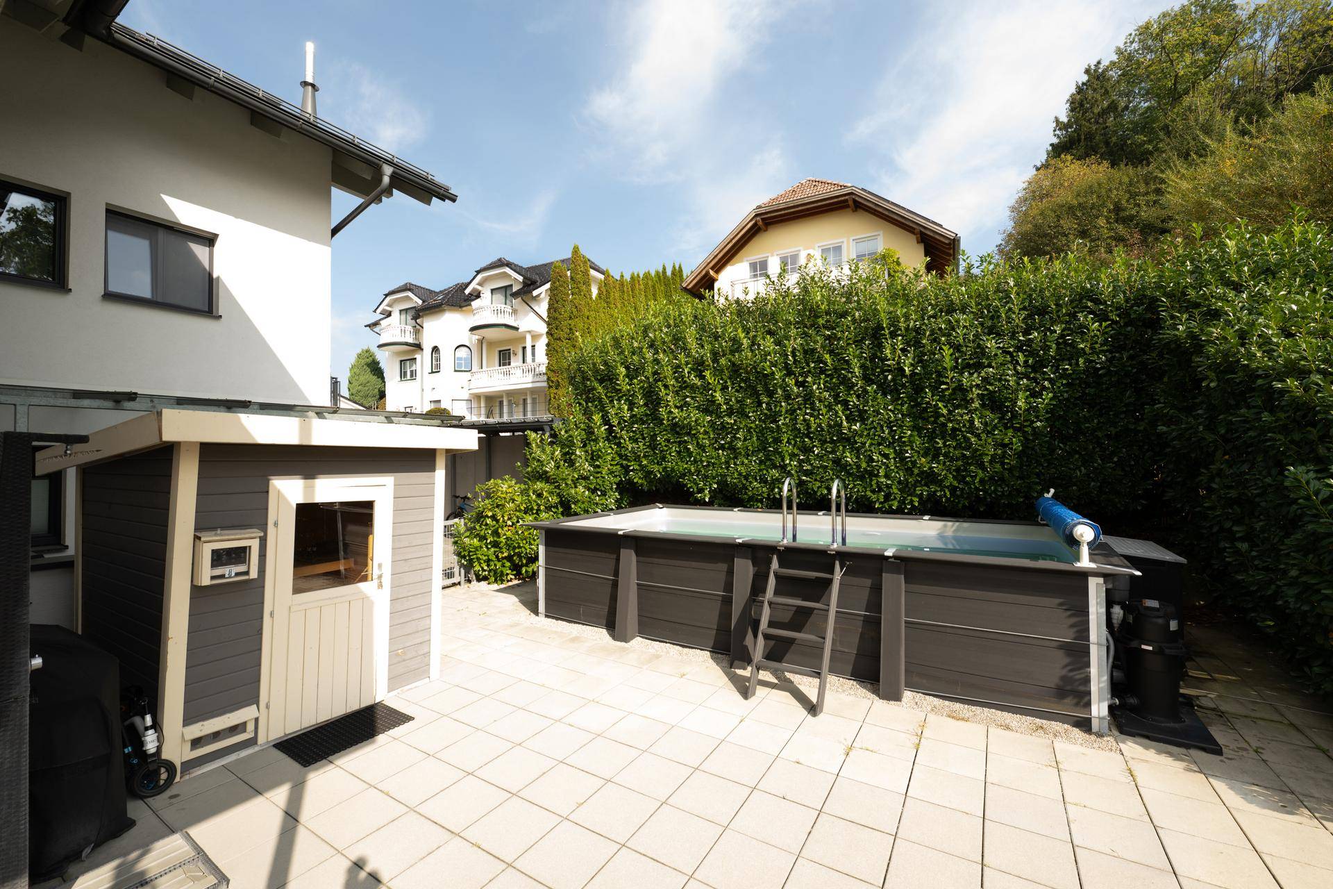 Gepflasterter Gartenbereich mit oberirdischem Pool, Gartenhaus und hoher Hecke.