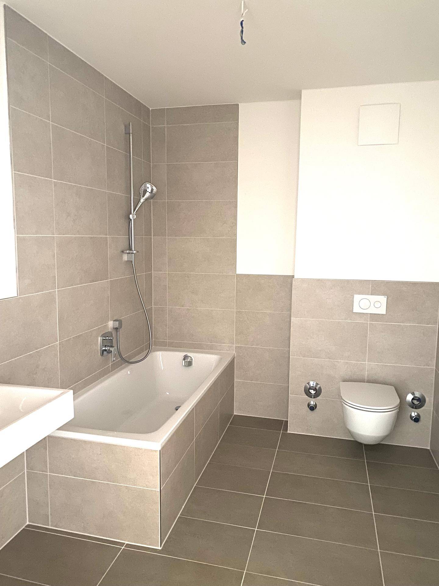 Modernes Badezimmer mit hellgrauen Fliesen, Badewanne mit Duschfunktion und wandhängendem WC.