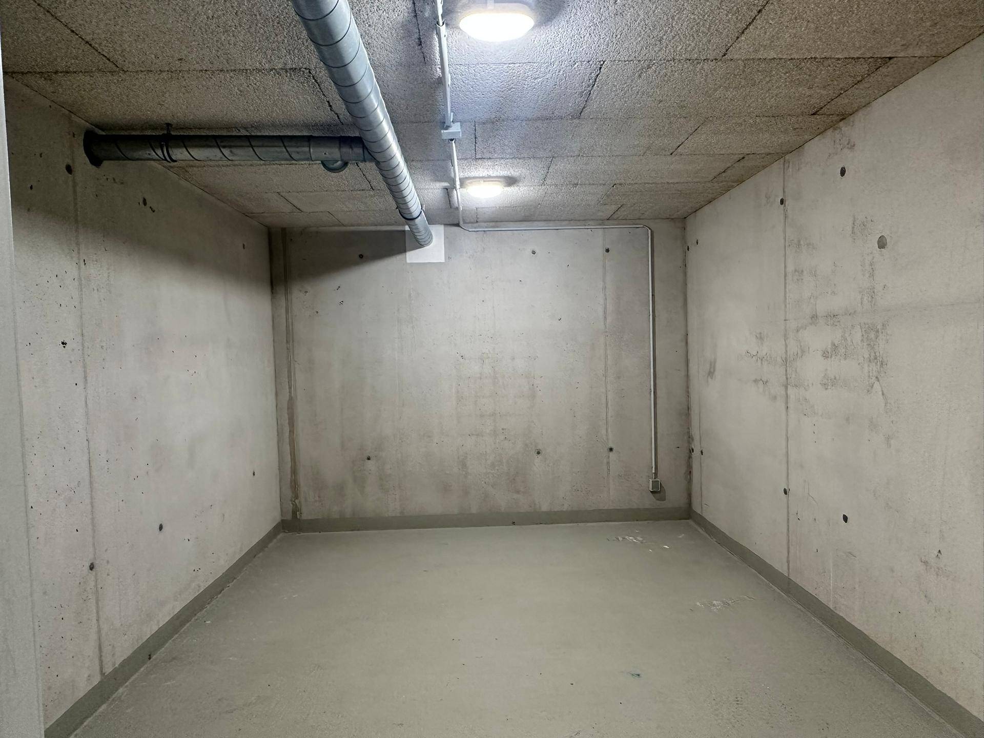 Leerer Abstellraum mit Sichtbetonwänden und Decke sowie sichtbaren Leitungen und Beleuchtung.