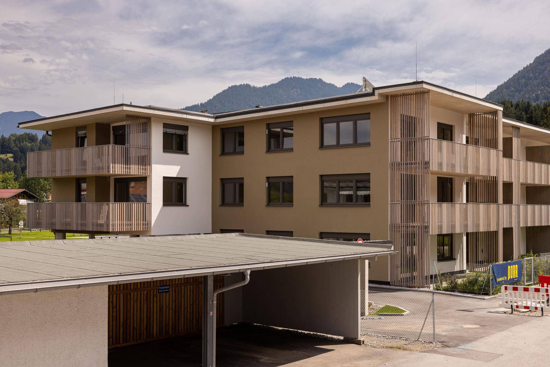 Modernes Mehrfamilienhaus mit Balkonen, Holzverkleidung und Carport, umgeben von Berglandschaft.