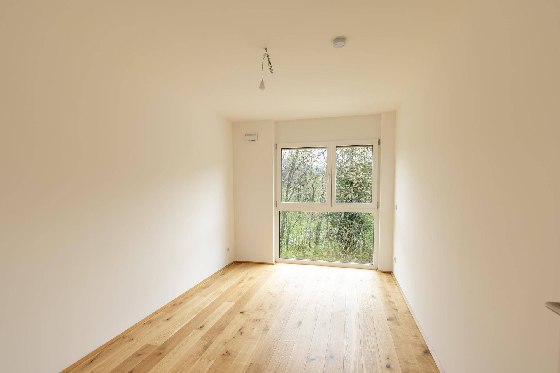 Helles Zimmer mit Holzboden und großem Fenster, das viel Tageslicht hereinlässt.