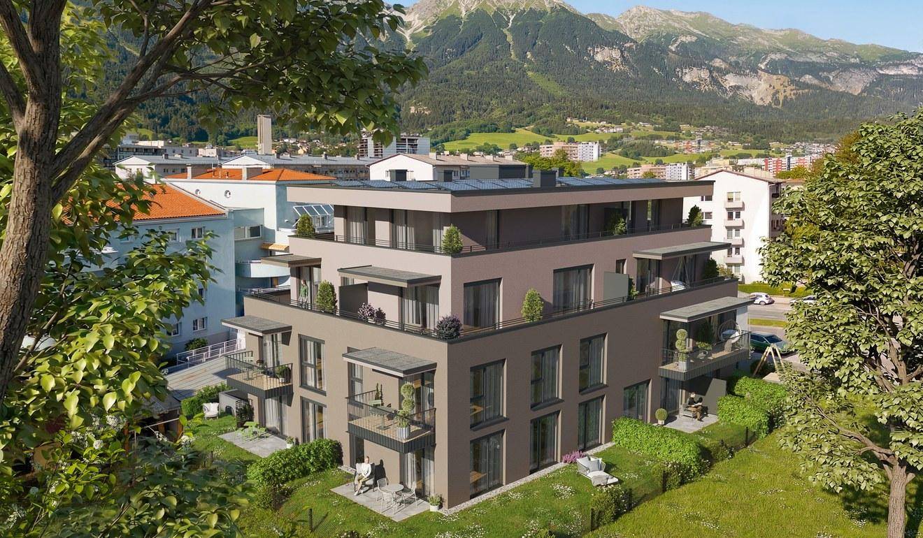 Luftaufnahme einer modernen Wohnanlage mit Flachdach, Balkonen und umliegender Grünfläche vor Bergkulisse.