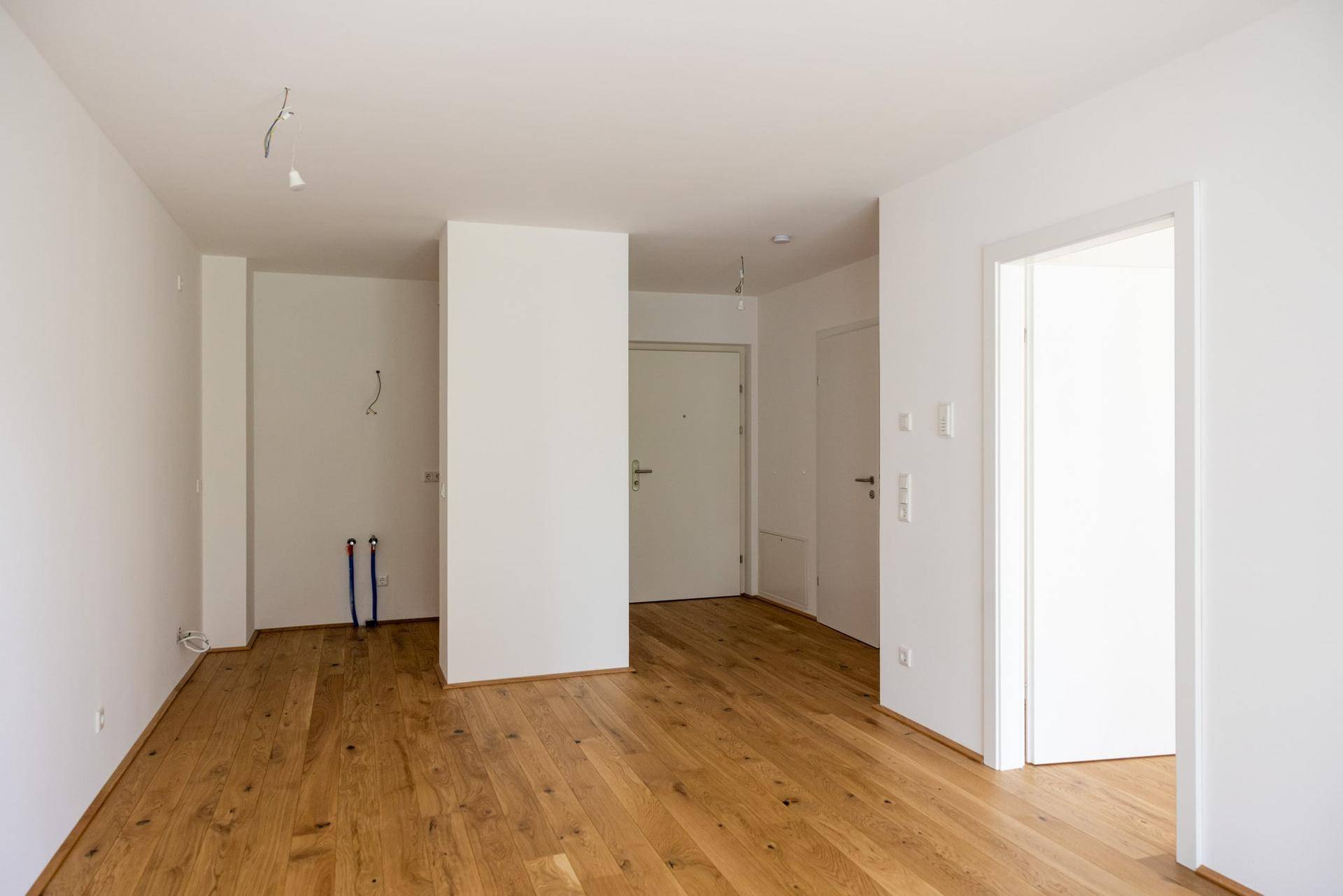 Heller Küchenbereich mit Holzboden, Installationsanschlüssen und Zugang zum Flur.