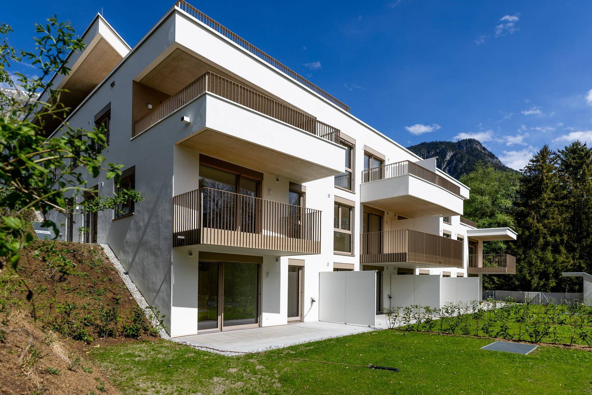 Neubau-Wohngebäude mit mehreren Balkonen und Terrassen in einer sonnigen, grünen Umgebung.