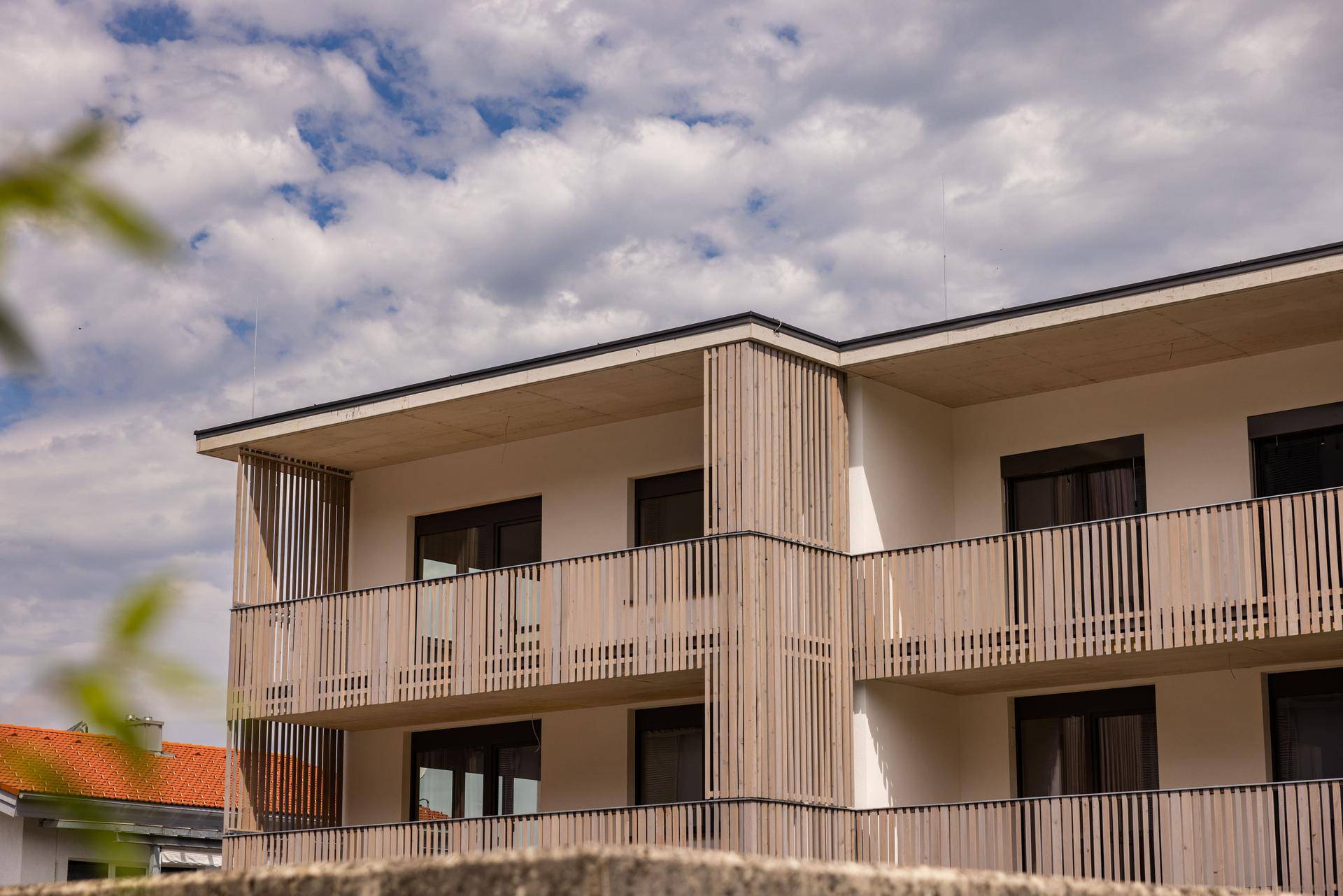 Moderne Gebäudefassade mit mehreren Balkonen und Holzlamellen-Verkleidung unter bewölktem Himmel.